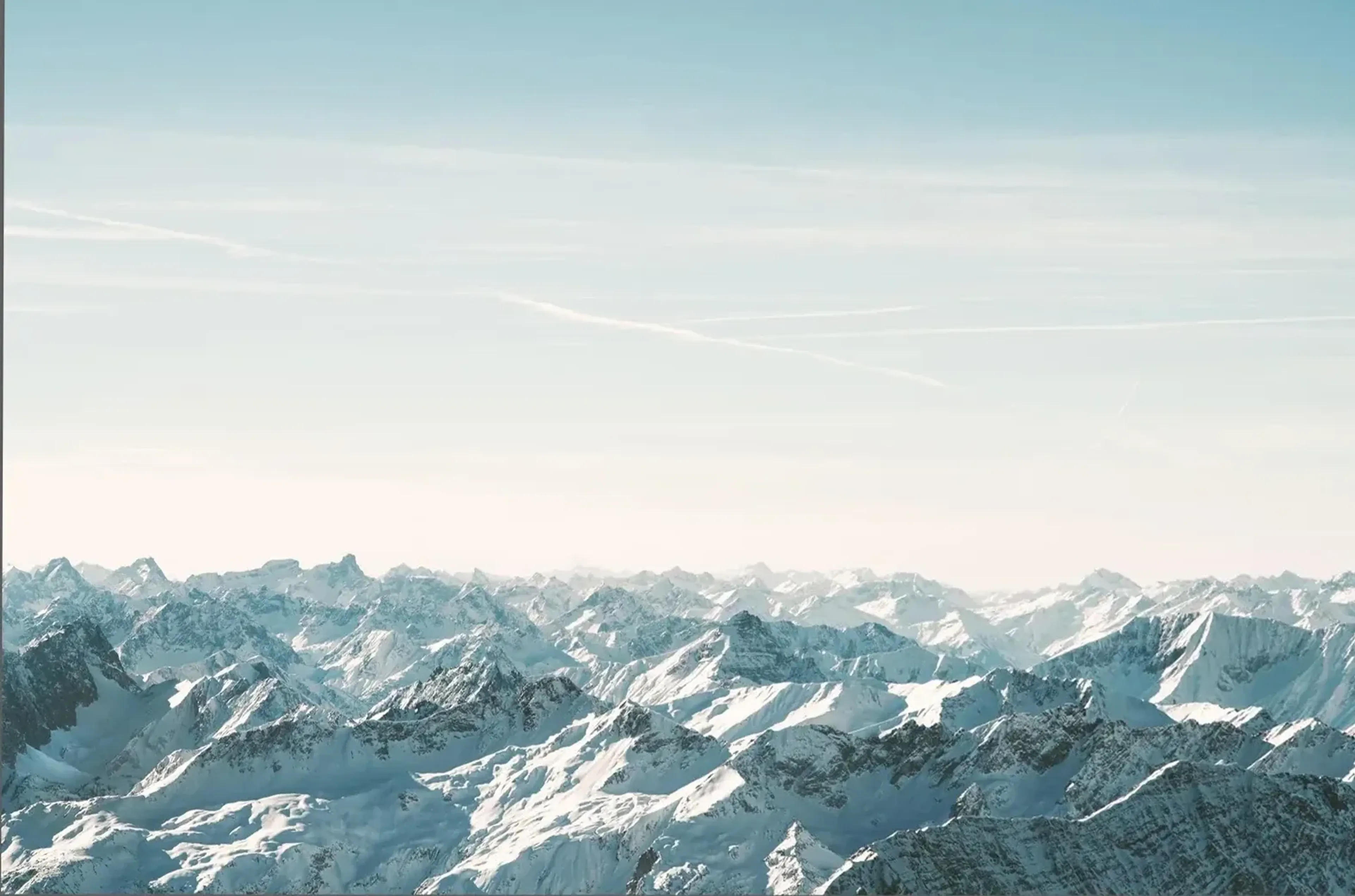 Schneebedeckte Bergketten reichen bis zum Horizont unter einem blassen Himmel mit feinen weißen Streifen.