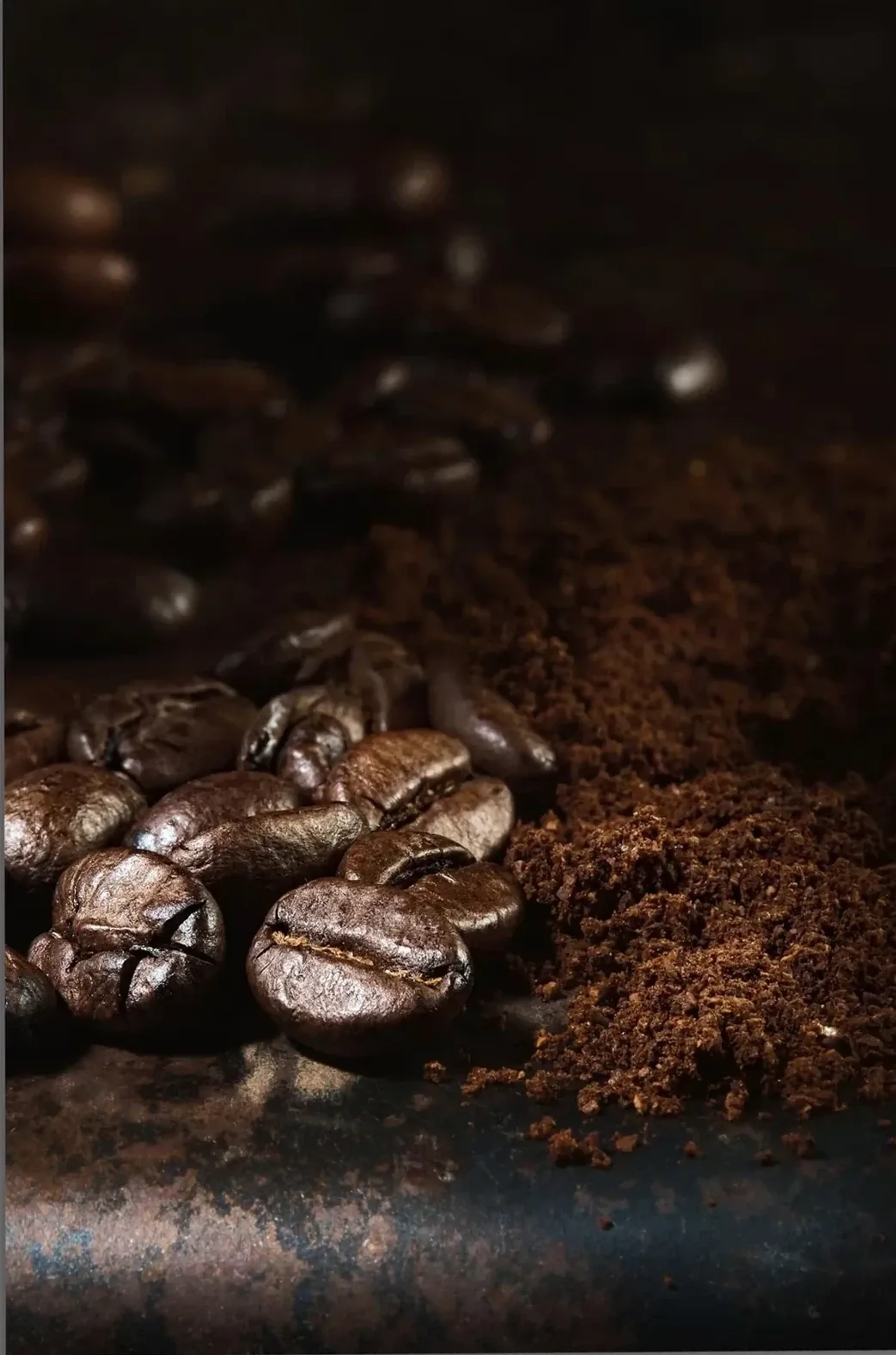 Nahaufnahme glänzender Kaffeebohnen neben feinem Kaffeepulver auf dunklem, gesprenkeltem Untergrund.