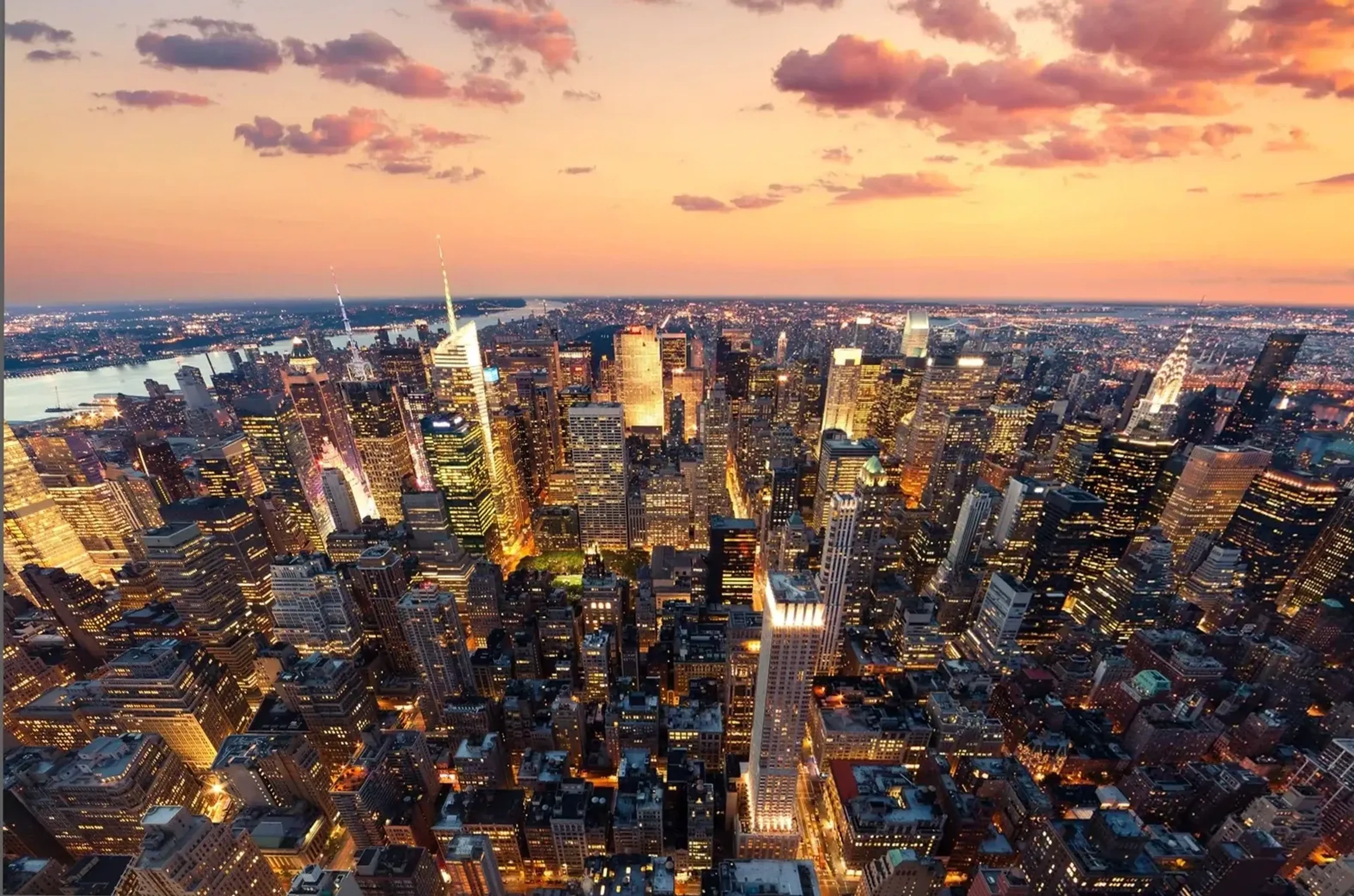 Manhattans Hochhäuser und leuchtende Straßenzüge aus hoher Perspektive unter einem pfirsichfarbenen Wolkenhimmel.