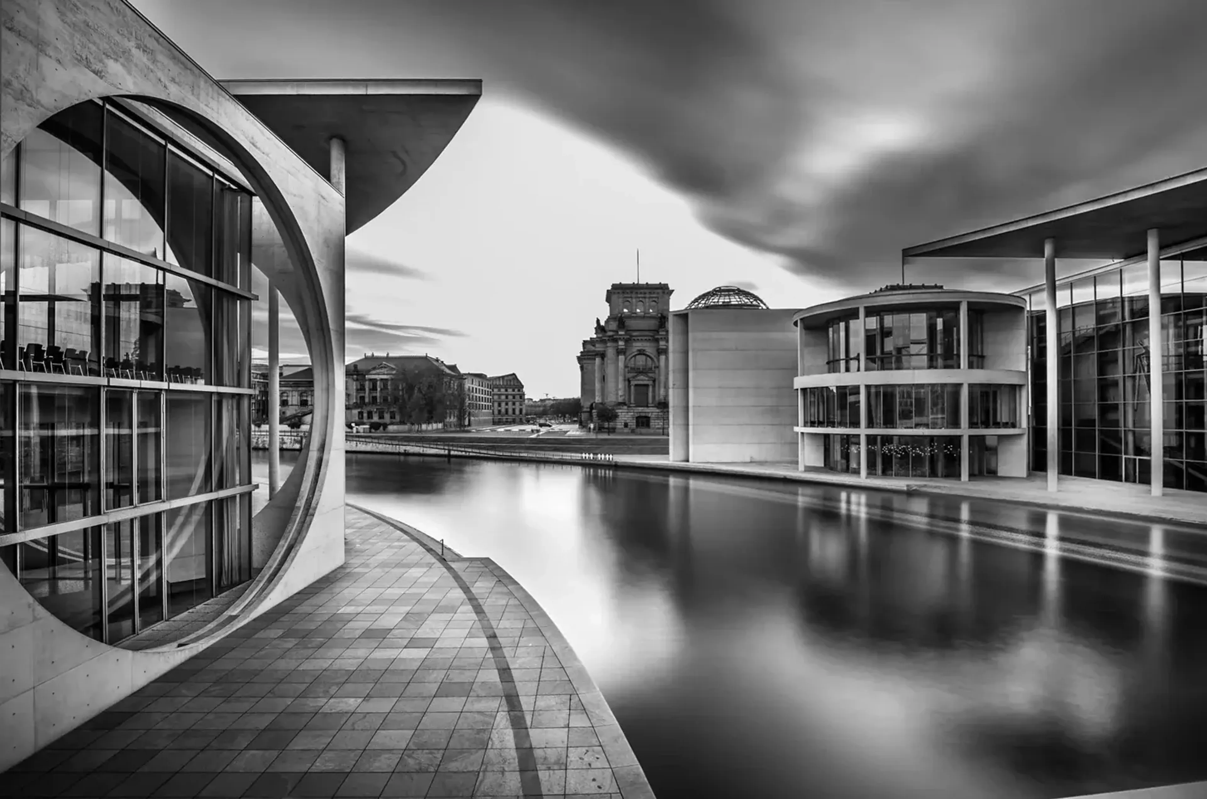 Schwarz-weiße Architektur am Wasser mit rundem Fassadenausschnitt, Spiegelungen und dunklen Wolken.