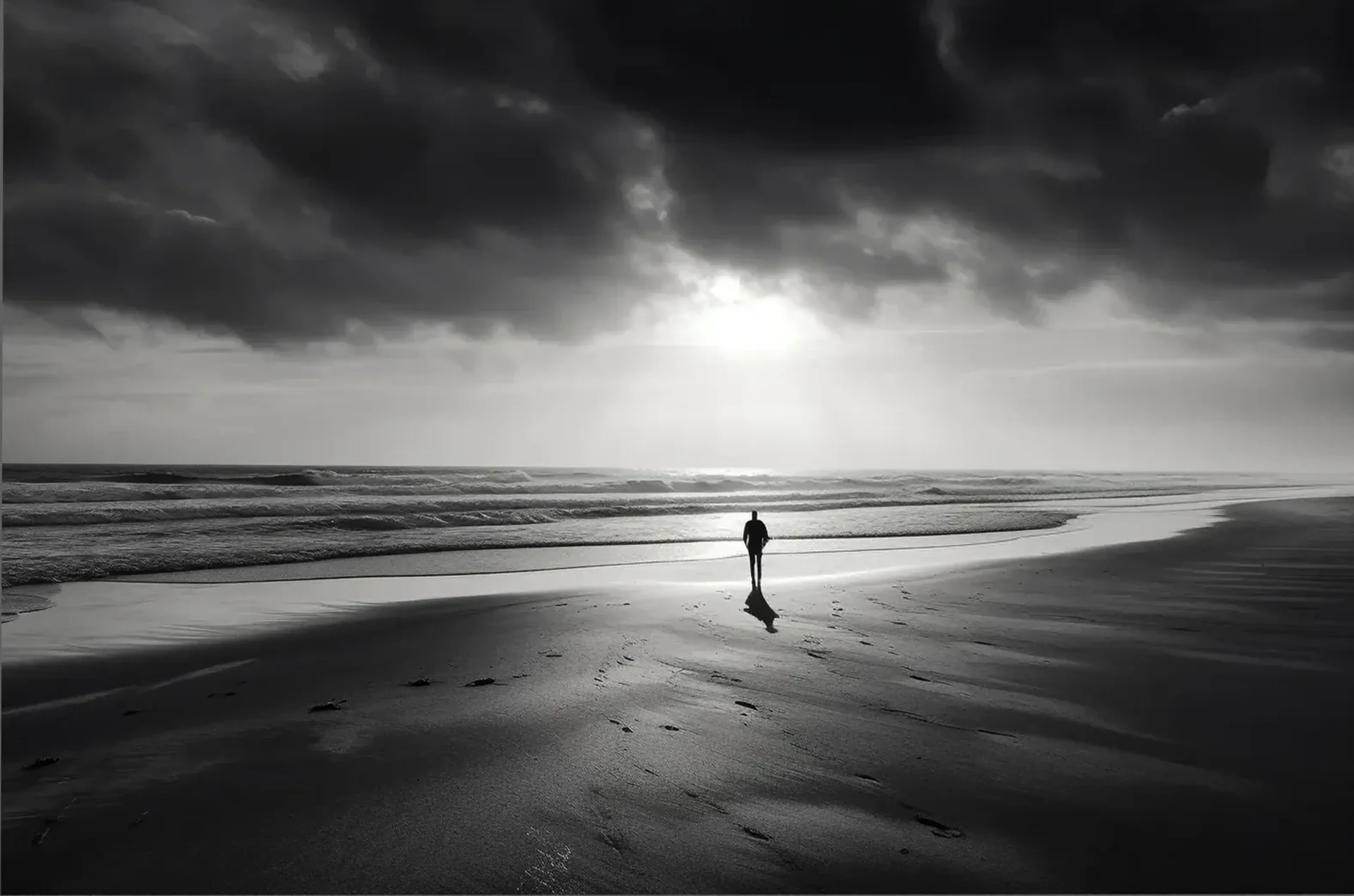 Eine einzelne Person am Strand unter dunklen Wolken und hellem Licht über dem Meer in Schwarz-Weiß.