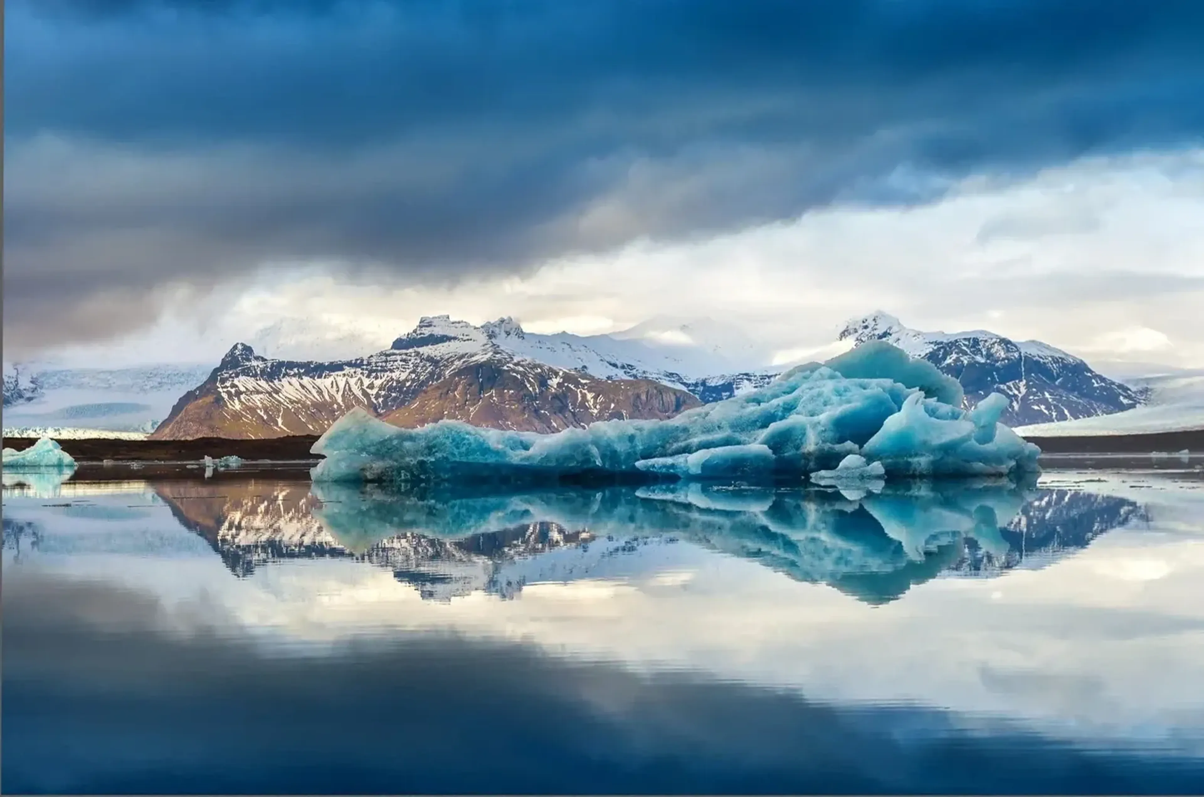 Ein türkisblauer Eisberg und schneebedeckte Berge spiegeln sich im Wasser unter dunklen Wolken.