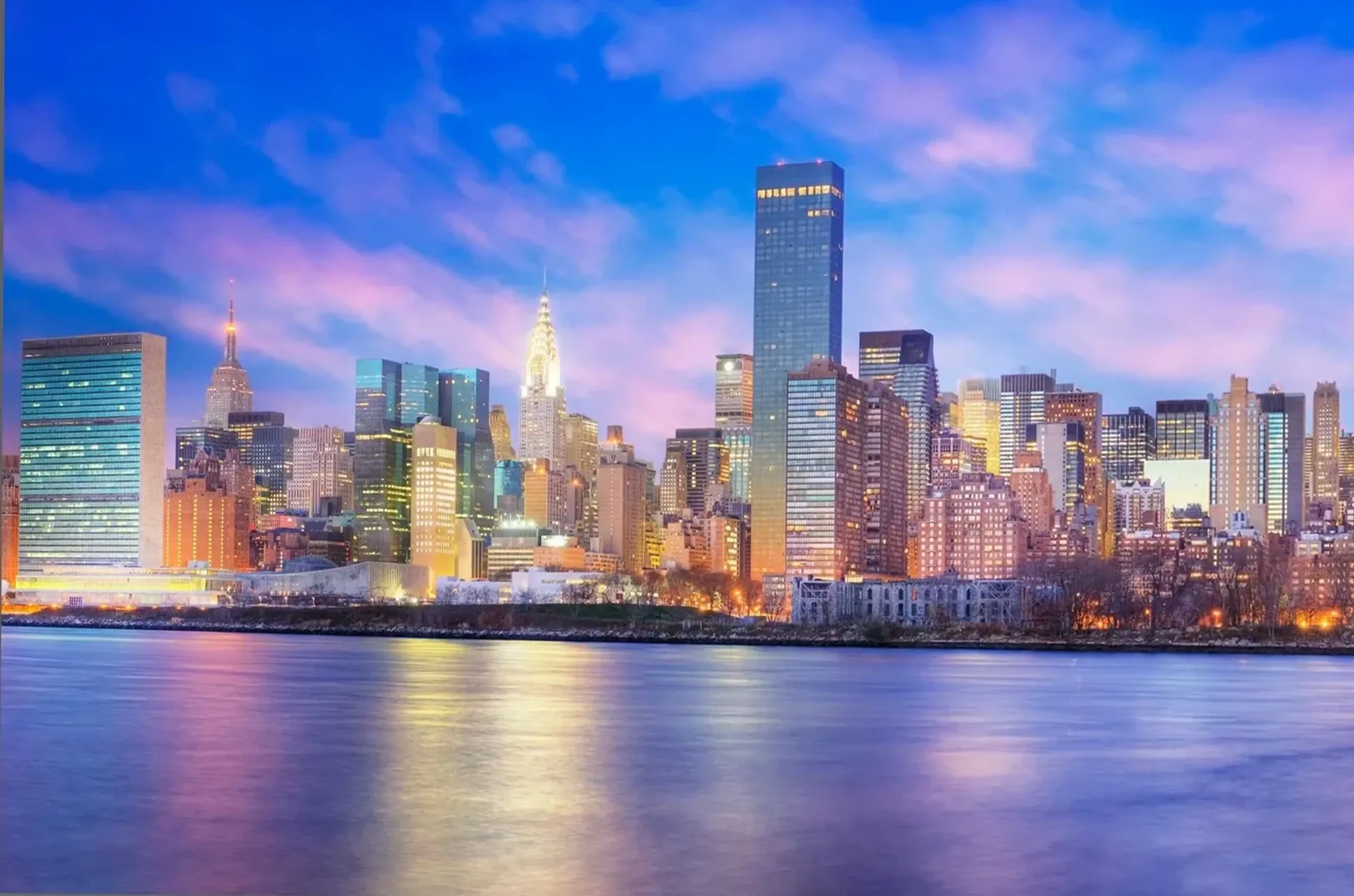 Manhattans beleuchtete Hochhäuser mit Chrysler Building hinter einer Wasserfläche unter blau-rosa Wolken.