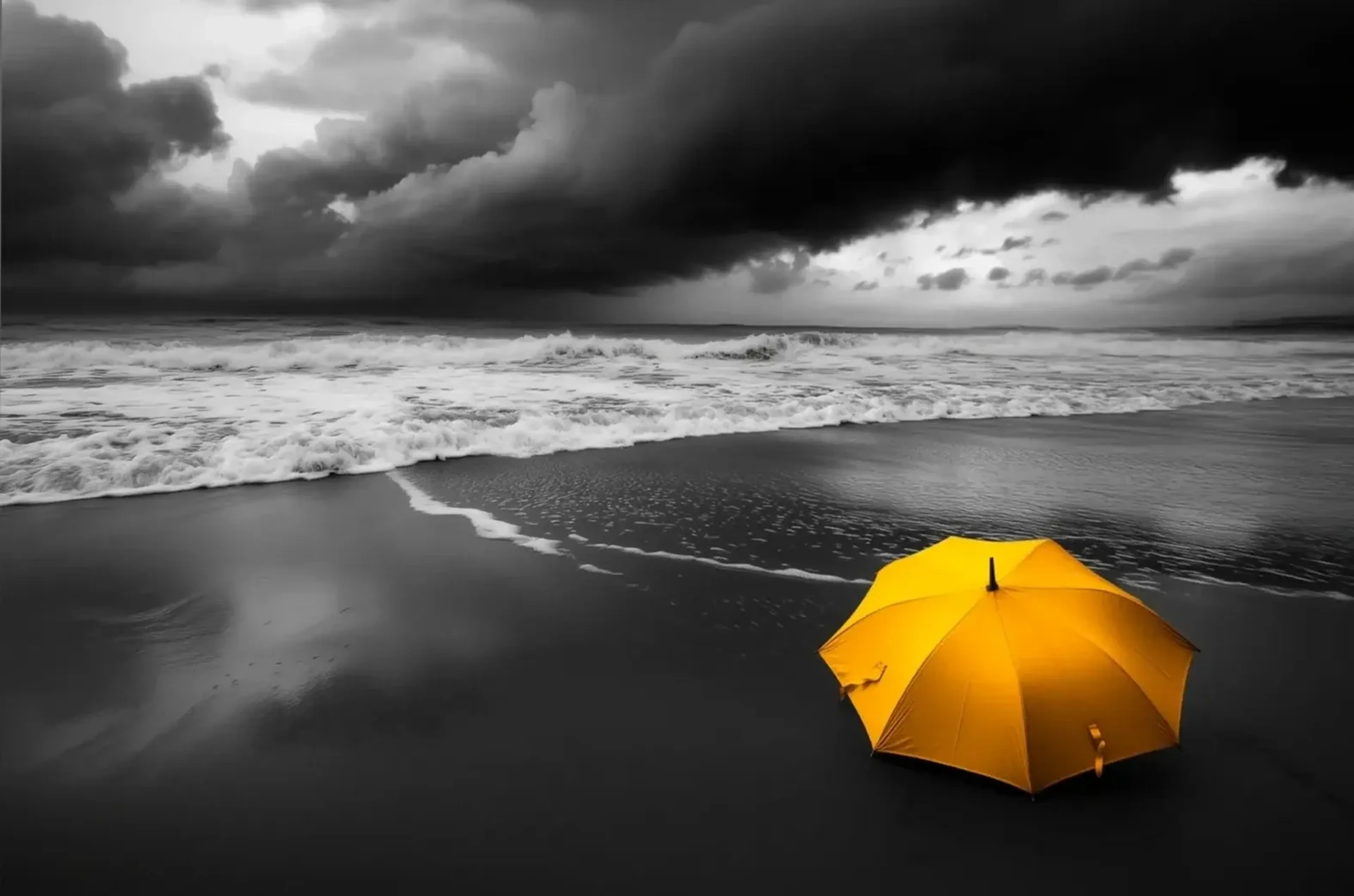 Ein gelber Regenschirm liegt am dunklen Strand vor schäumenden Wellen und schweren Wolken in Schwarz-Weiß.