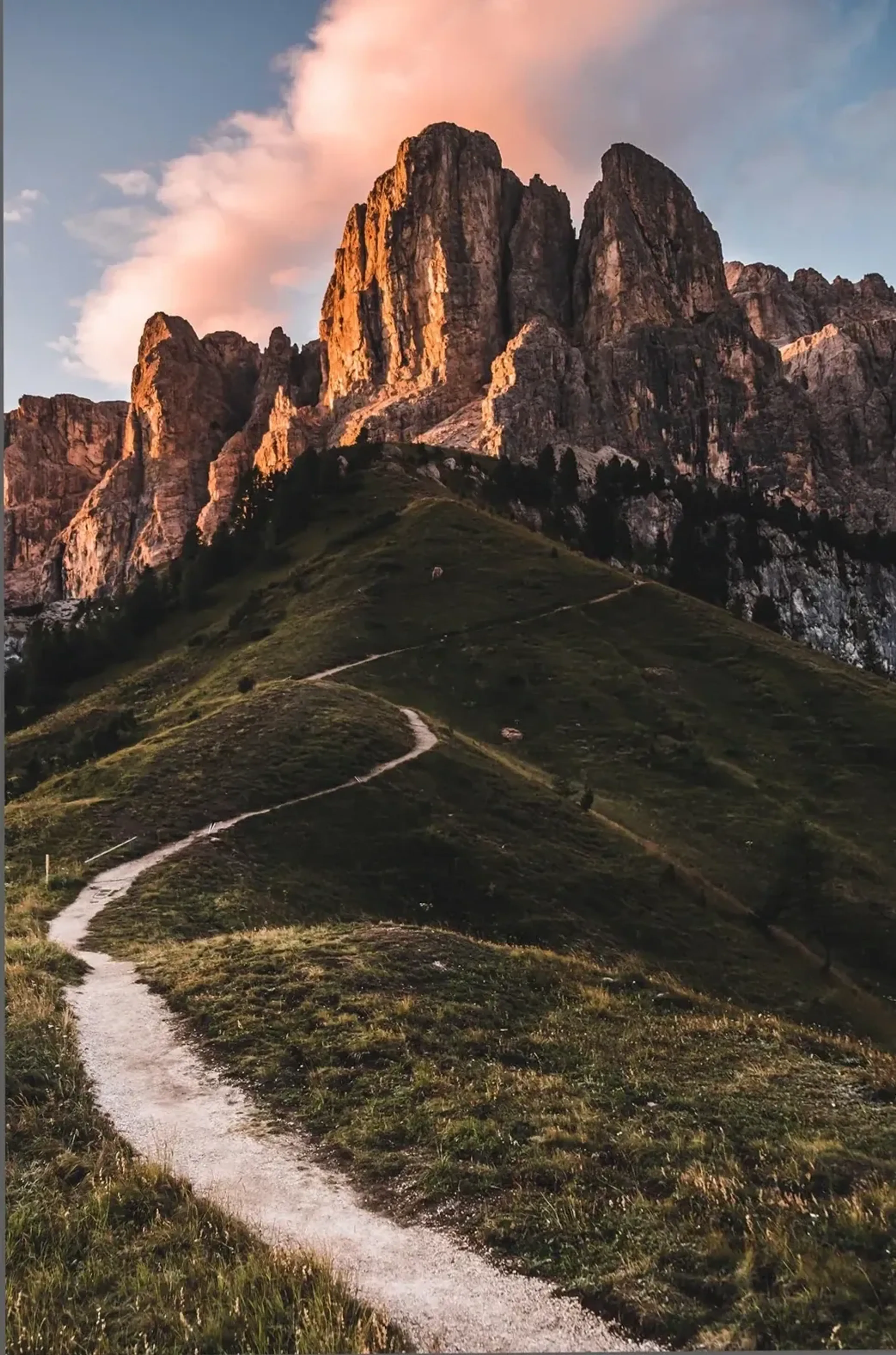 Ein heller Wanderpfad windet sich über grüne Hänge zu orange beleuchteten Felstürmen unter rosa Wolken.