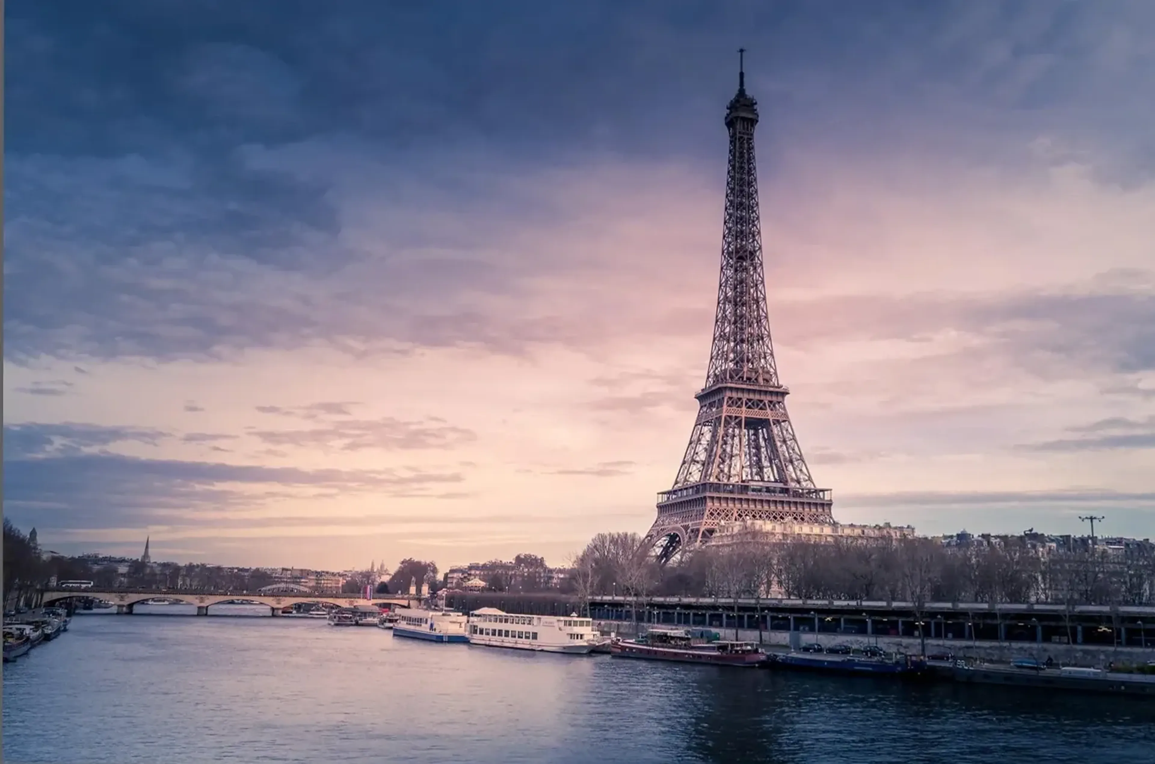 Der Eiffelturm steht an der Seine mit Booten am Ufer unter einem blaugrauen und rosafarbenen Himmel.