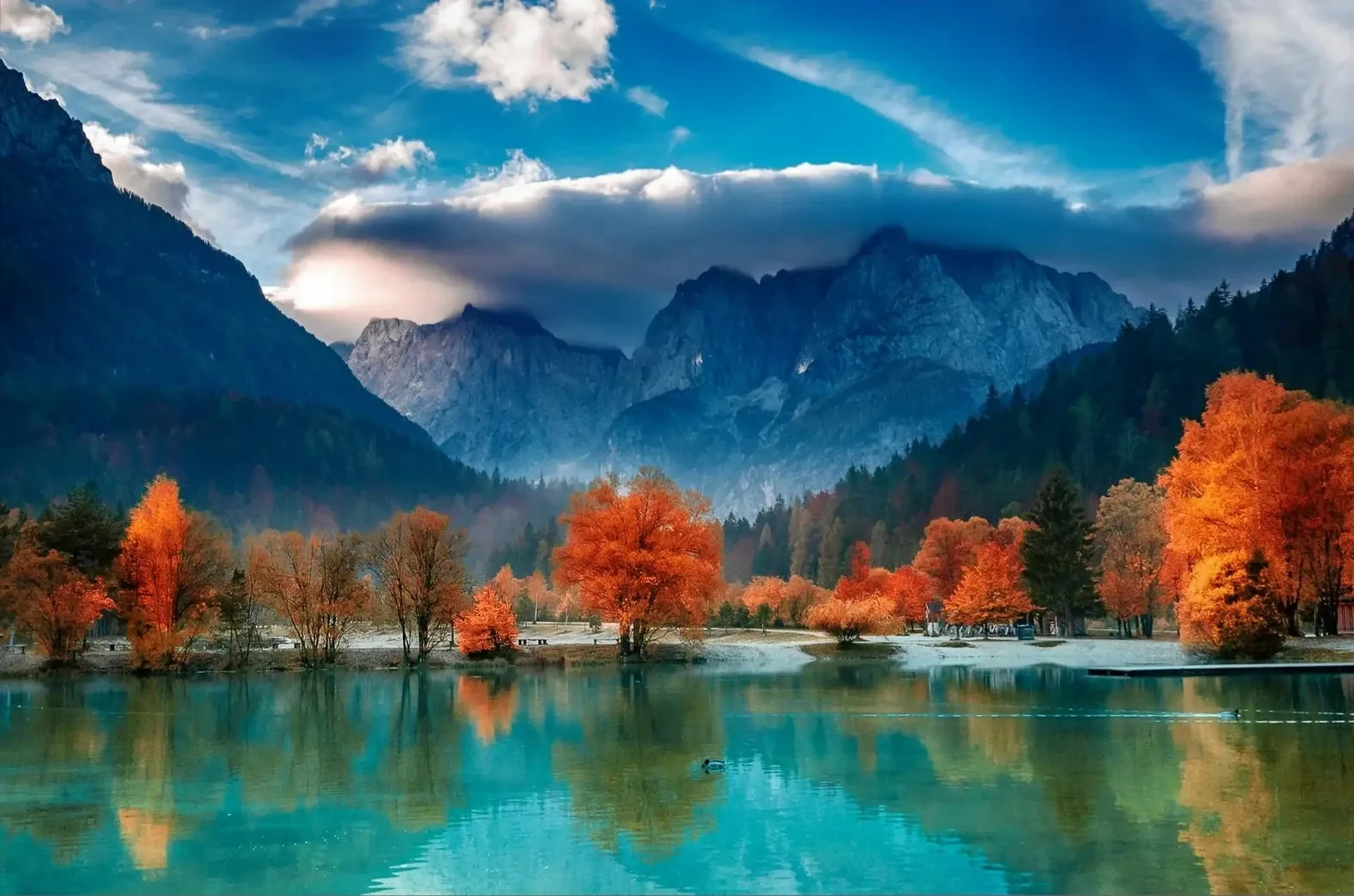 Orangefarbene Herbstbäume spiegeln sich in einem türkisfarbenen See vor felsigen Bergen und tiefen Wolken.