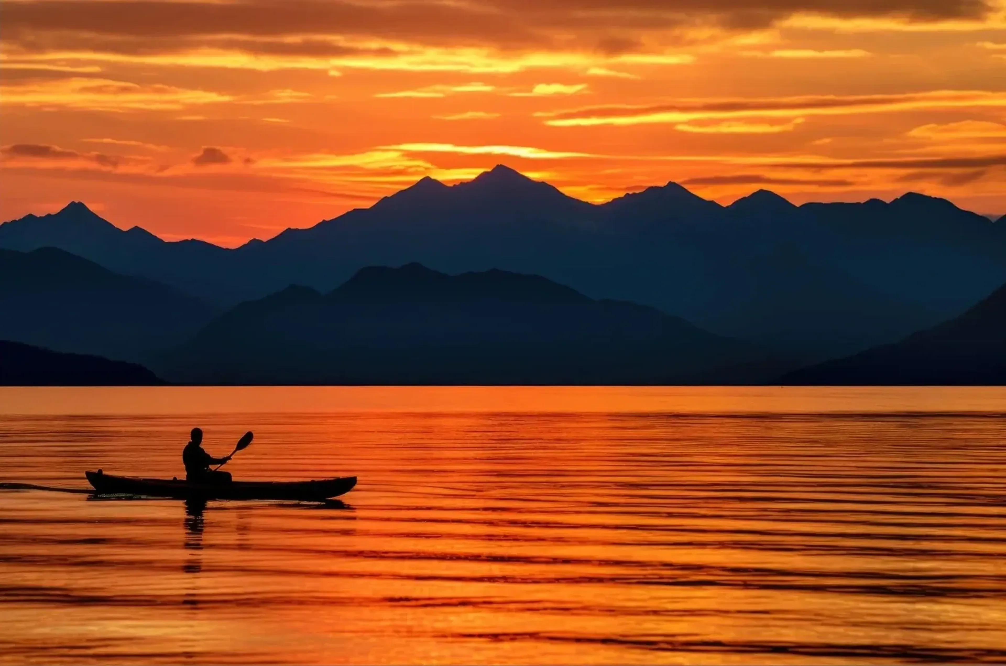 Ein Paddler im Kanu fährt über orange schimmerndes Wasser vor dunklen Bergketten im Abendlicht.