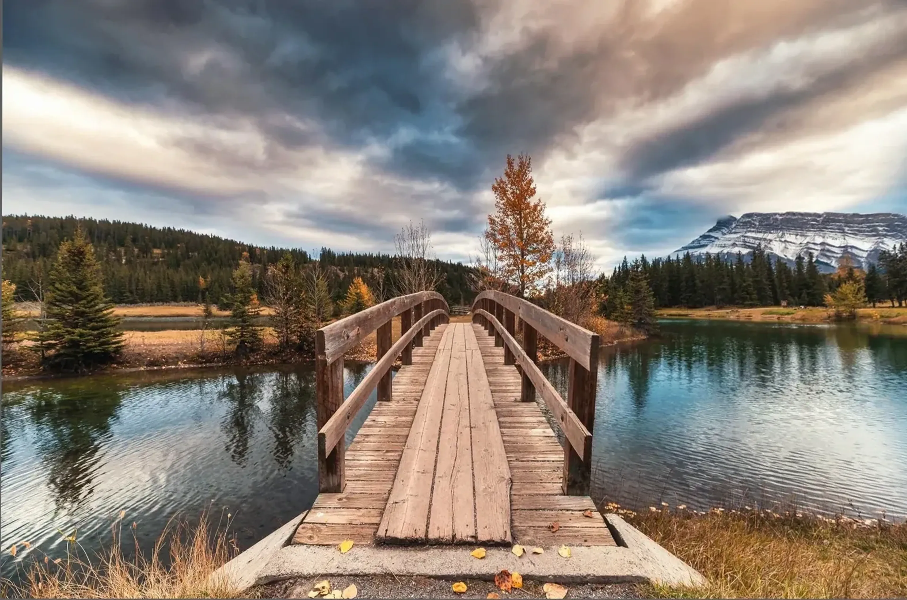 Eine Holzbrücke führt über einen See zu herbstlichen Bäumen, dahinter liegen Wald und ein Berg unter Wolken.