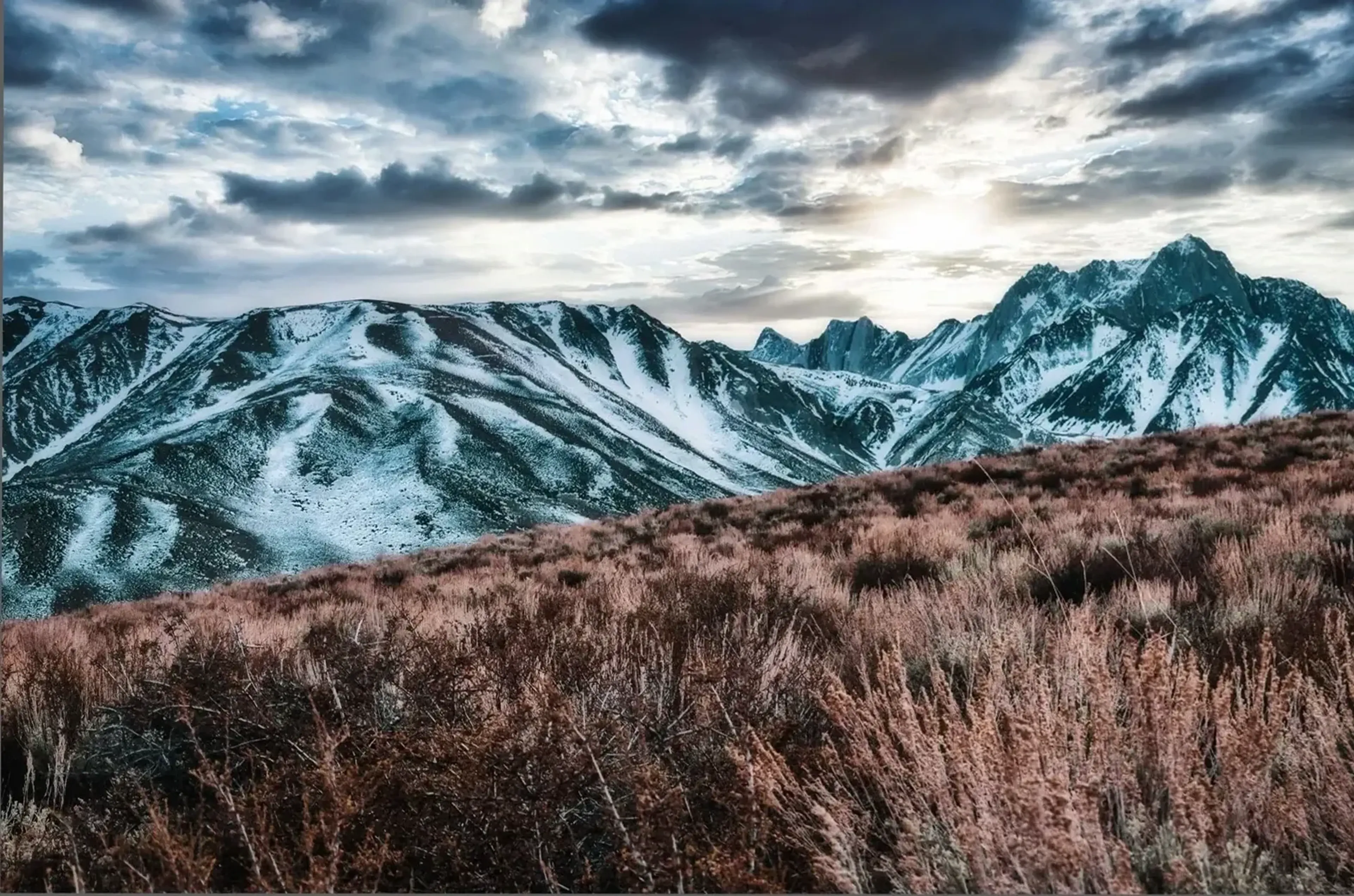 Rosabraune Gräser liegen vor schneegestreiften Bergkämmen, über denen Licht durch eine dichte Wolkendecke fällt.