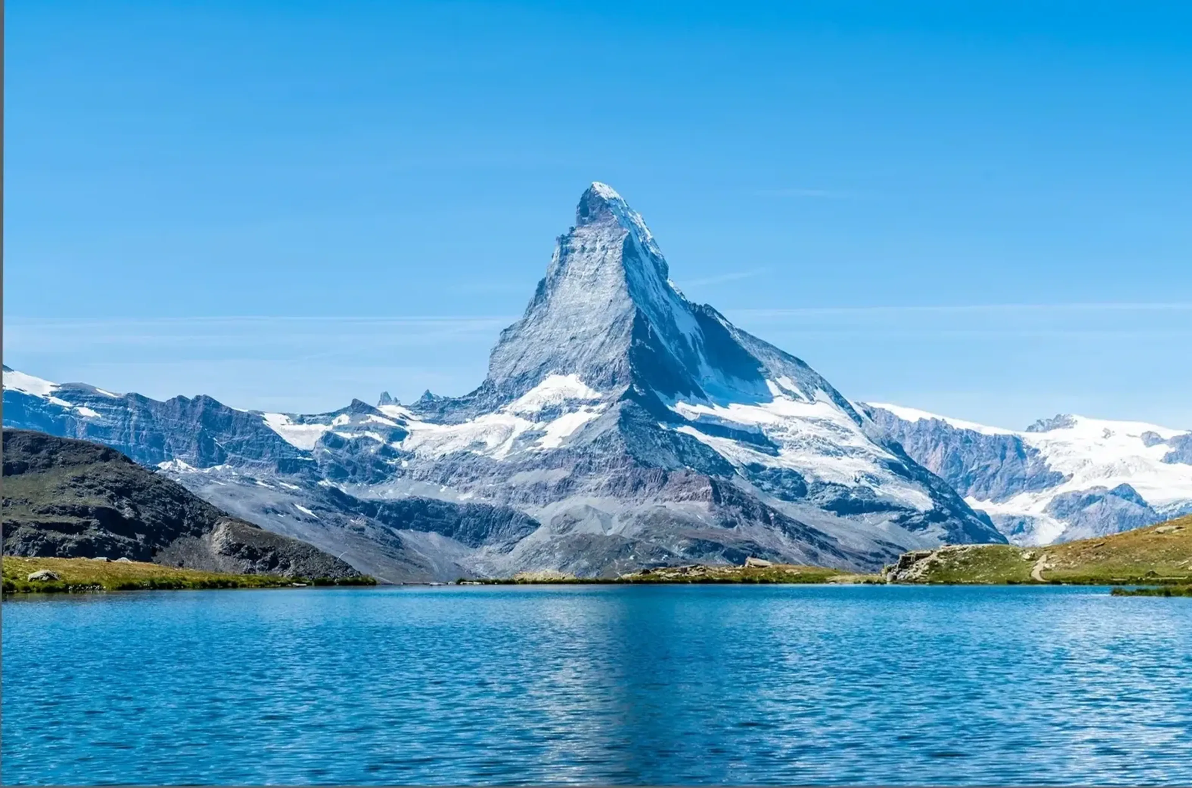 Ein spitzer, teilweise schneebedeckter Berg erhebt sich hinter einem türkisblauen See unter klarem Himmel.