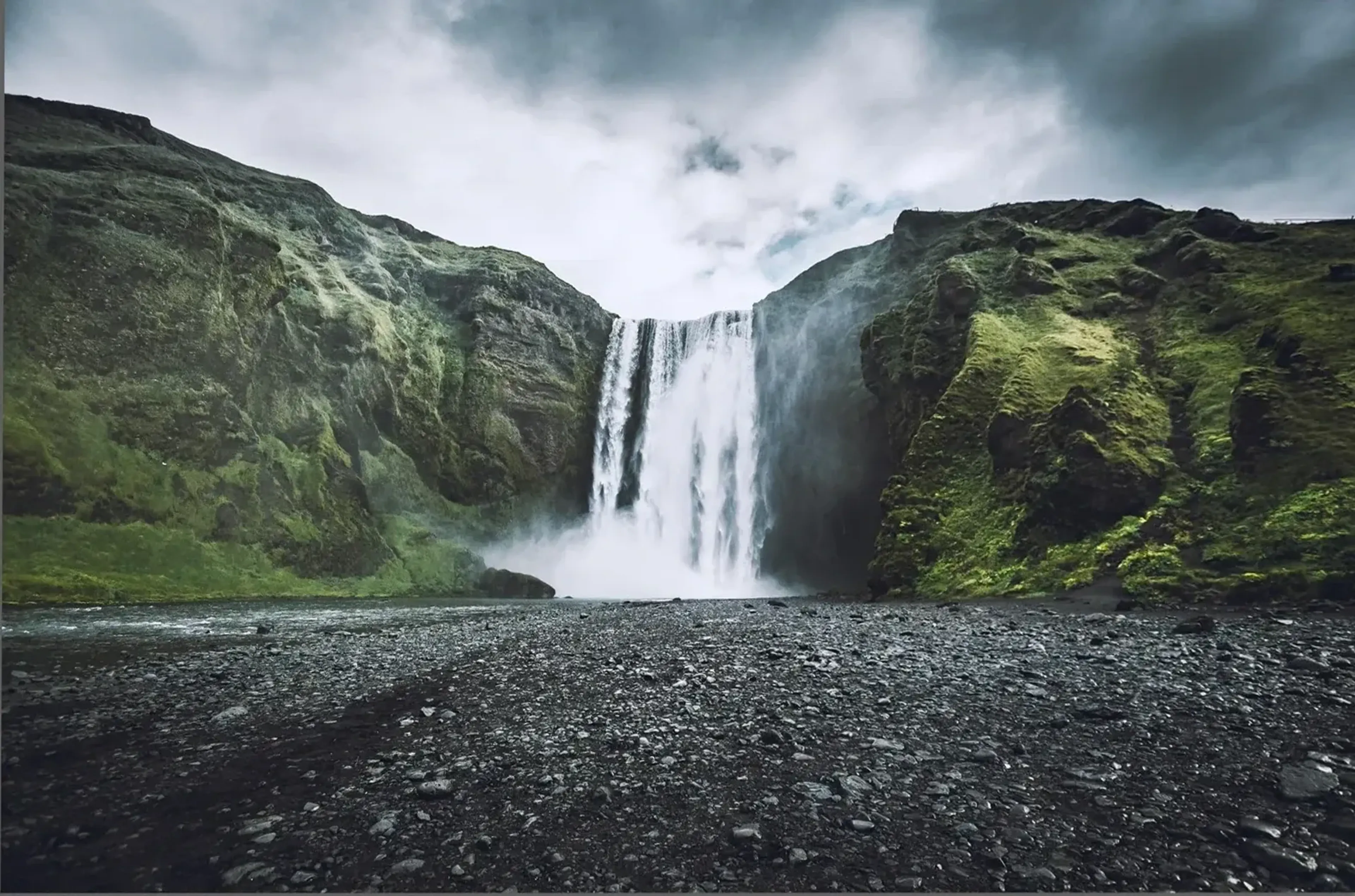 Breiter Wasserfall zwischen grünen Felswänden, davor dunkler Kies und darüber ein grauer Wolkenhimmel.