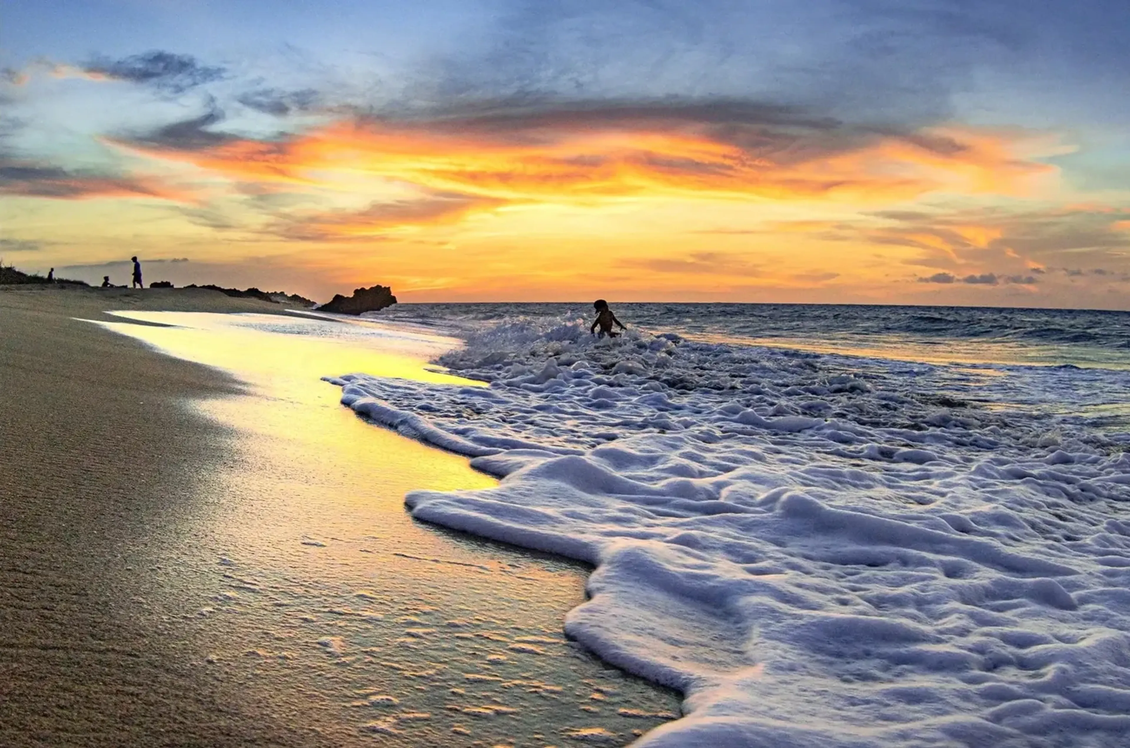 Weißer Wellenschaum am Sandstrand unter einem orangegelben Abendhimmel, mit kleinen Menschenfiguren am Wasser.
