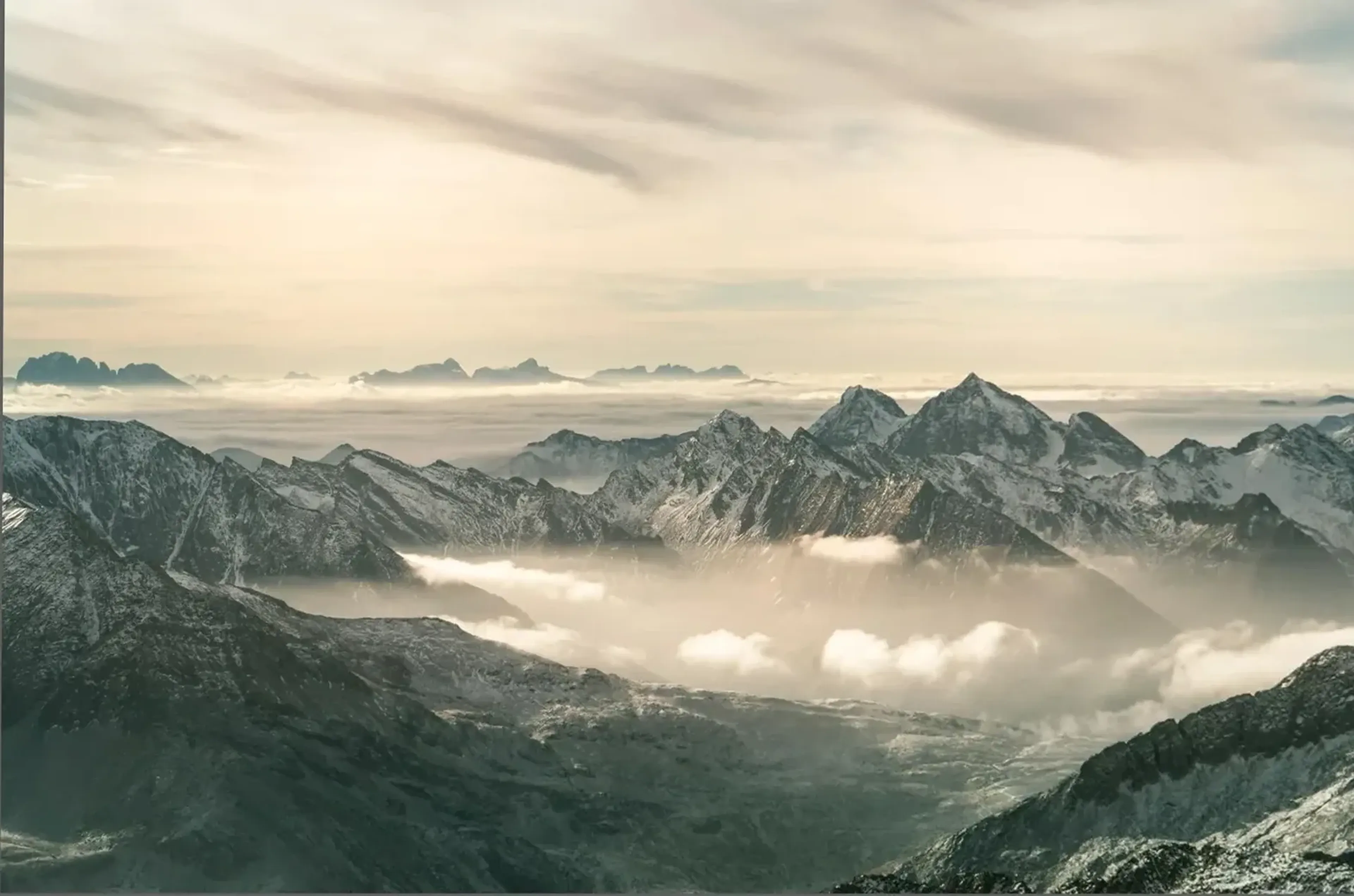 Schneebedeckte Bergketten ragen zwischen tief liegenden Wolken unter einem hellen, cremefarbenen Himmel auf.