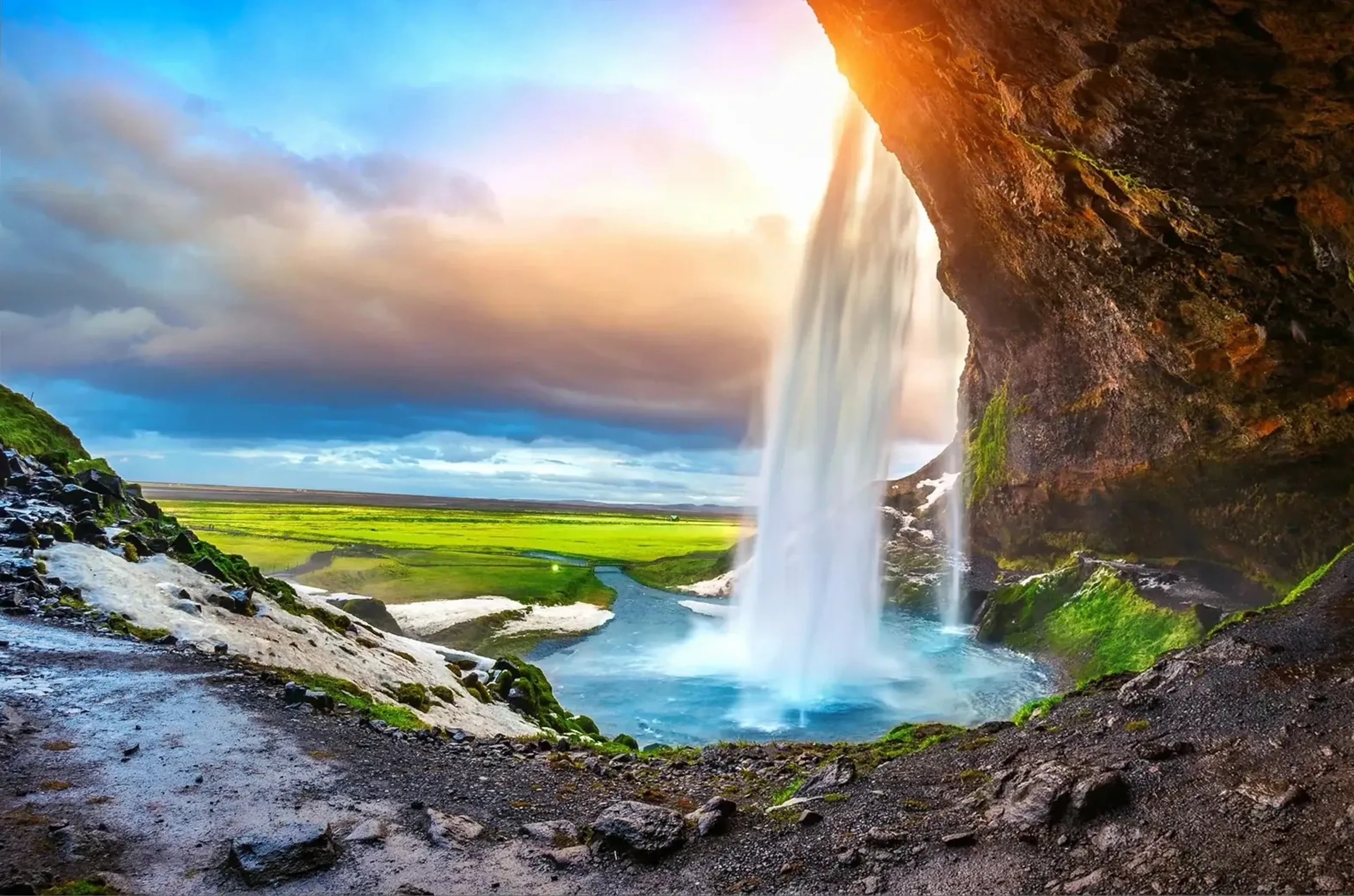 Blick unter einem Felsüberhang auf einen Wasserfall, ein türkisfarbenes Becken und eine grüne Ebene im warmen Licht.