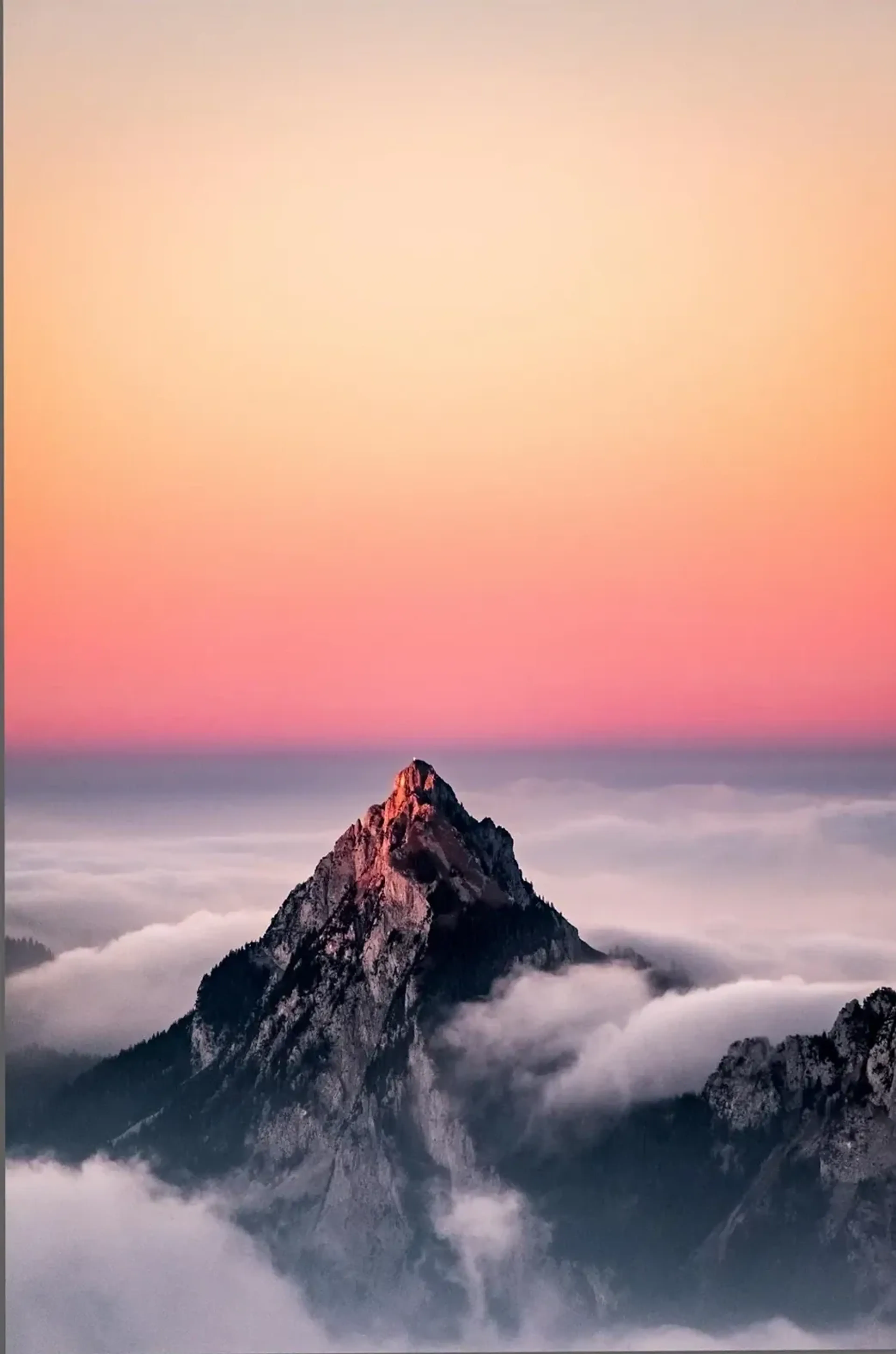 Ein schroffer Berggipfel ragt aus Wolken unter einem Himmel in Rosa und Pfirsichtönen.