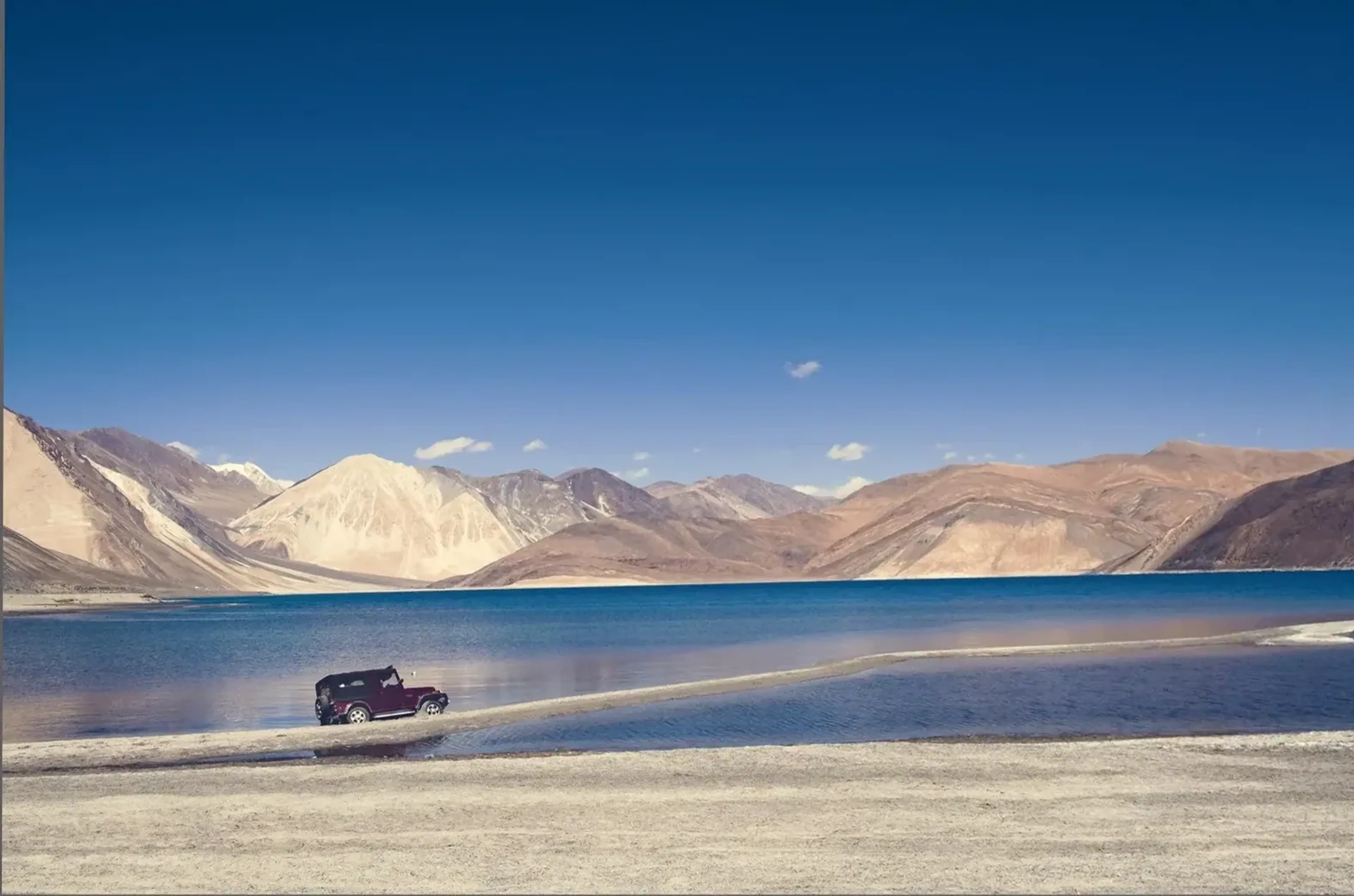 Ein dunkler Geländewagen steht am blauen See vor kahlen, sandfarbenen Bergen unter klarem Himmel.