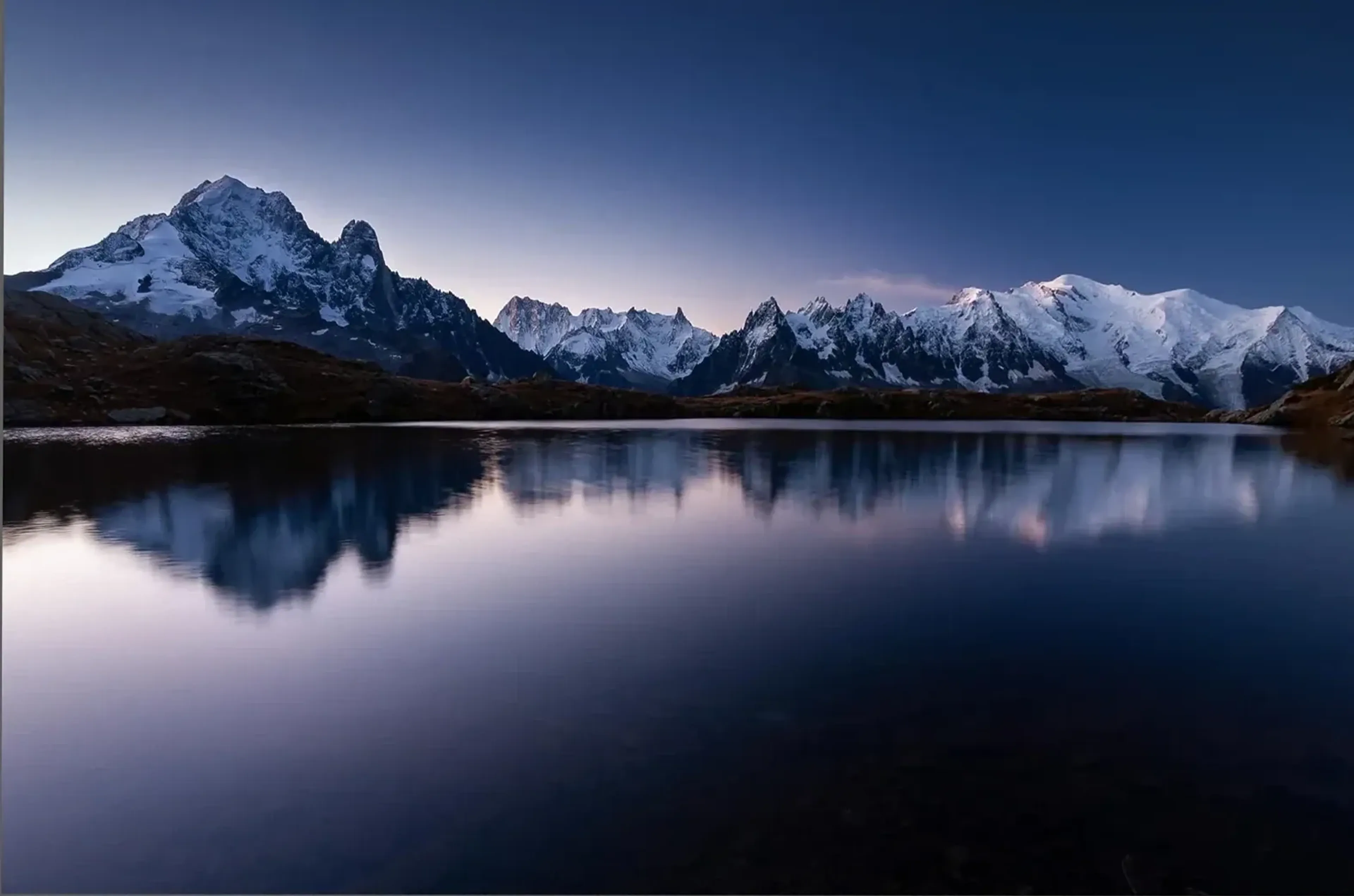 Eine schneebedeckte Bergkette spiegelt sich in einem stillen See unter einem dunkelblauen Himmel.
