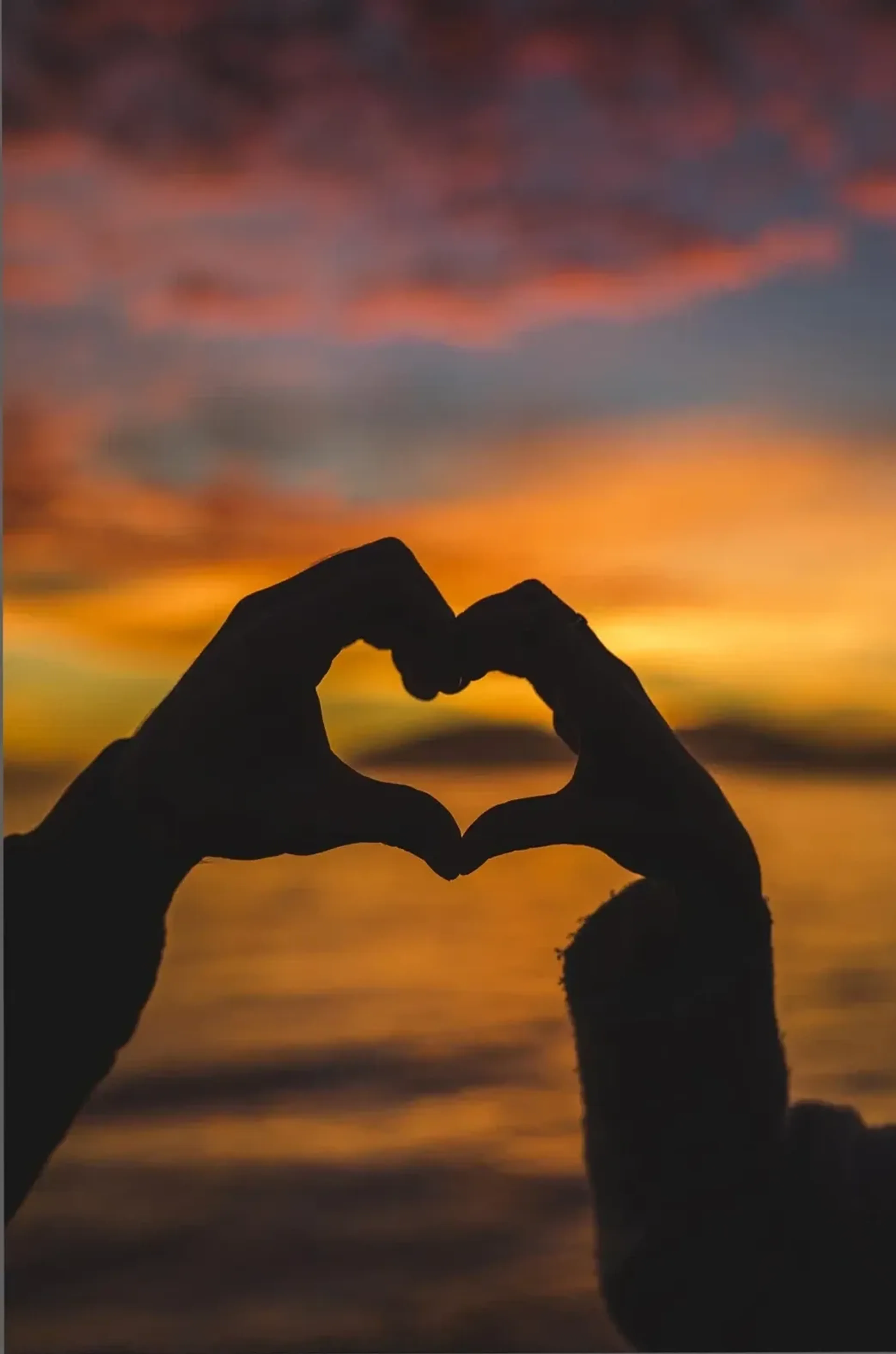 Zwei Hände formen eine dunkle Herzsilhouette vor einem orangegoldenen Sonnenuntergang über dem Wasser.