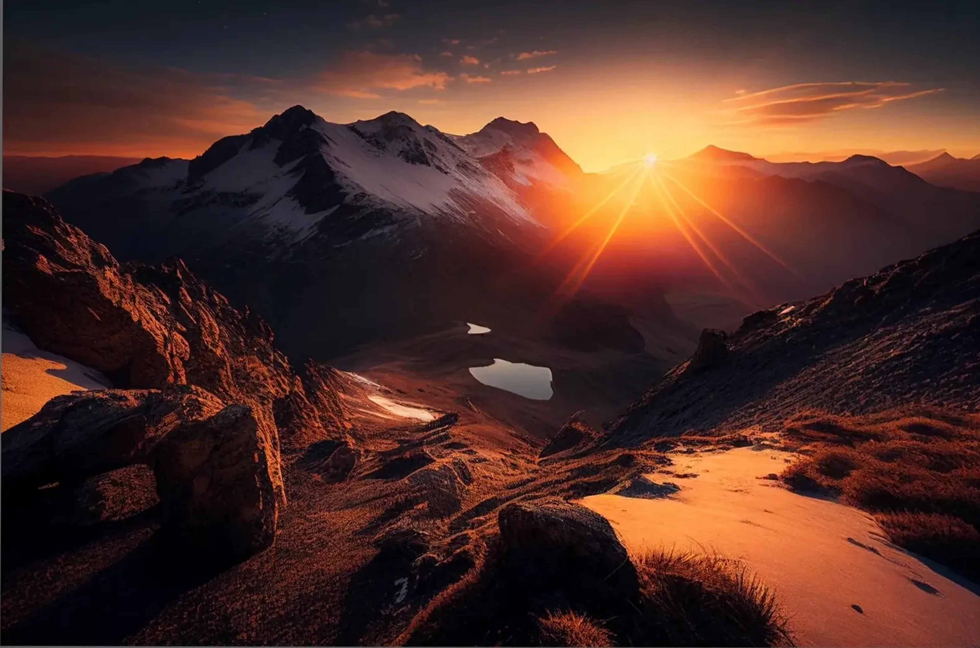 Orangefarbener Sonnenuntergang über einem Bergtal mit zwei kleinen Seen, dunklen Felsen und Schneefeldern.