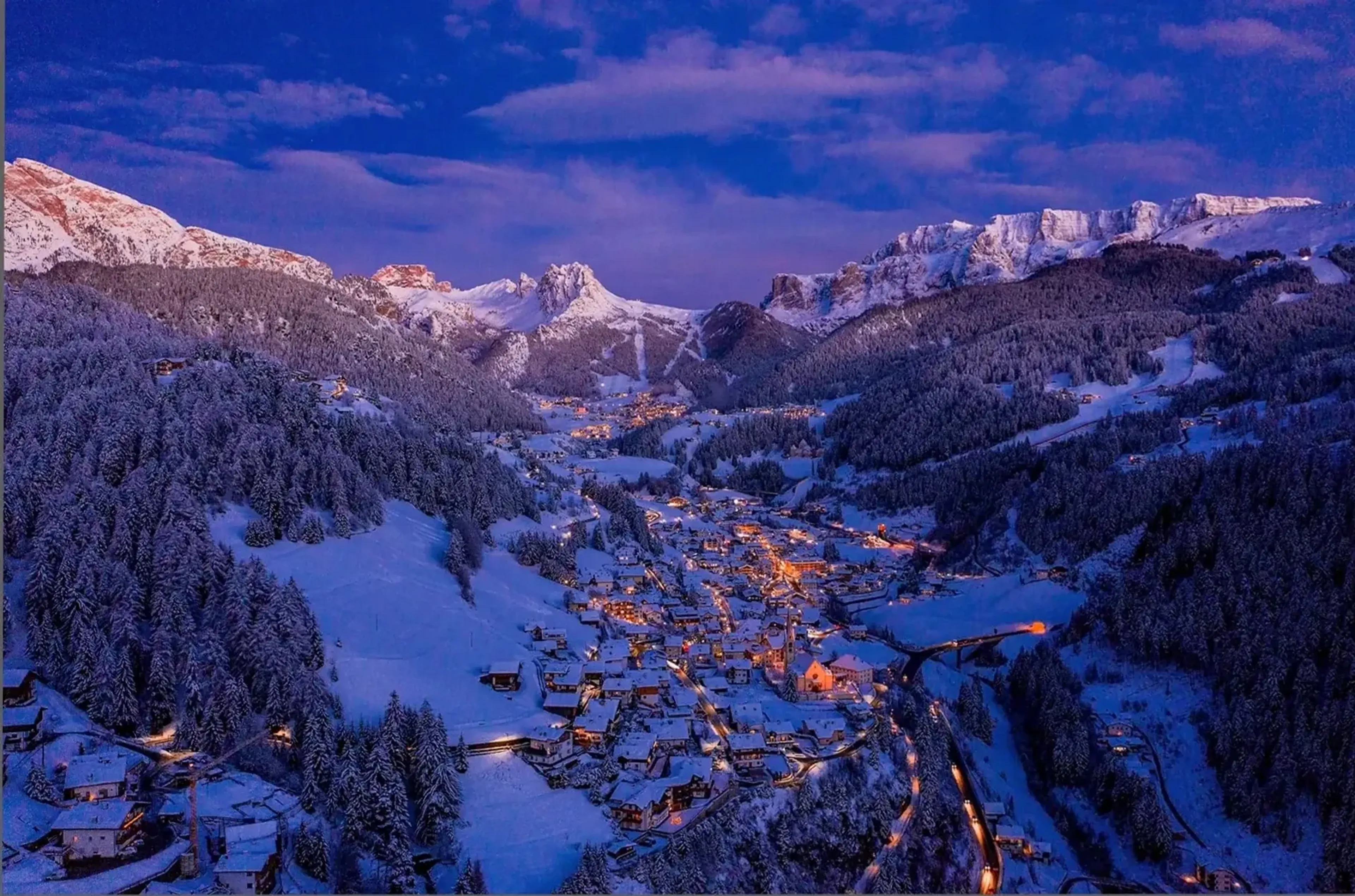 Warme Dorflichter leuchten in einem verschneiten Tal zwischen dunklen Wäldern und Bergen unter blauviolettem Himmel.