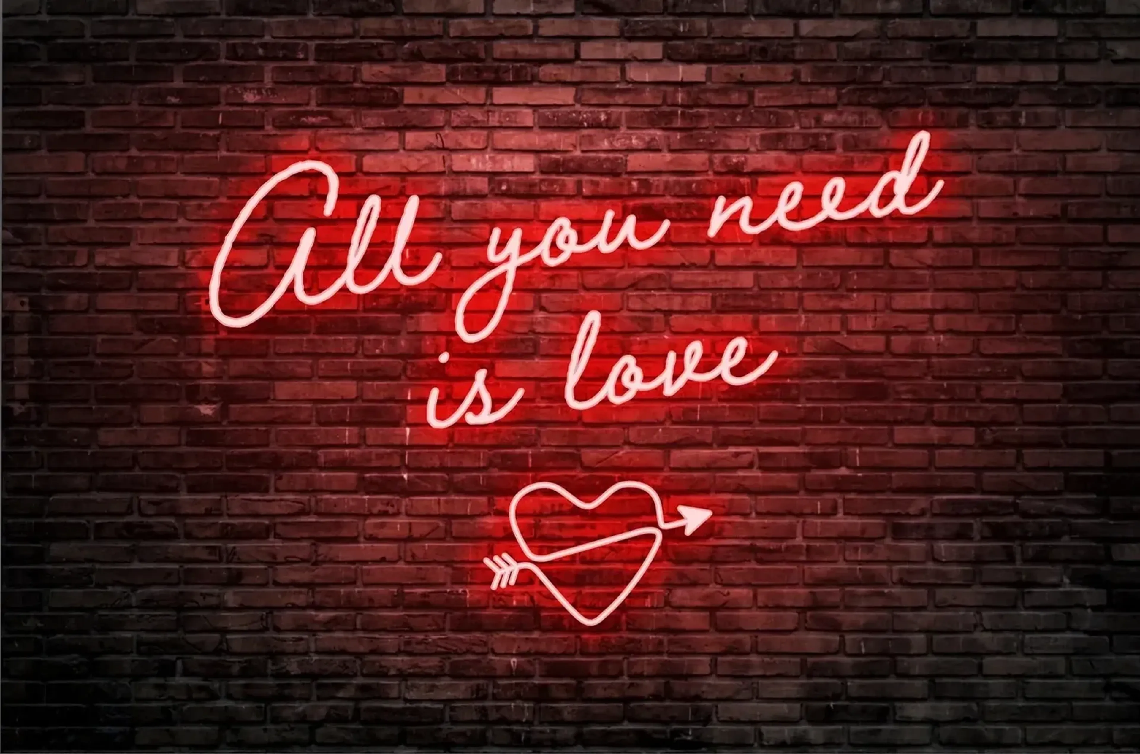 Der rote Neonschriftzug „All you need is love“ leuchtet mit einem Herz und Pfeil auf dunkler Backsteinwand.