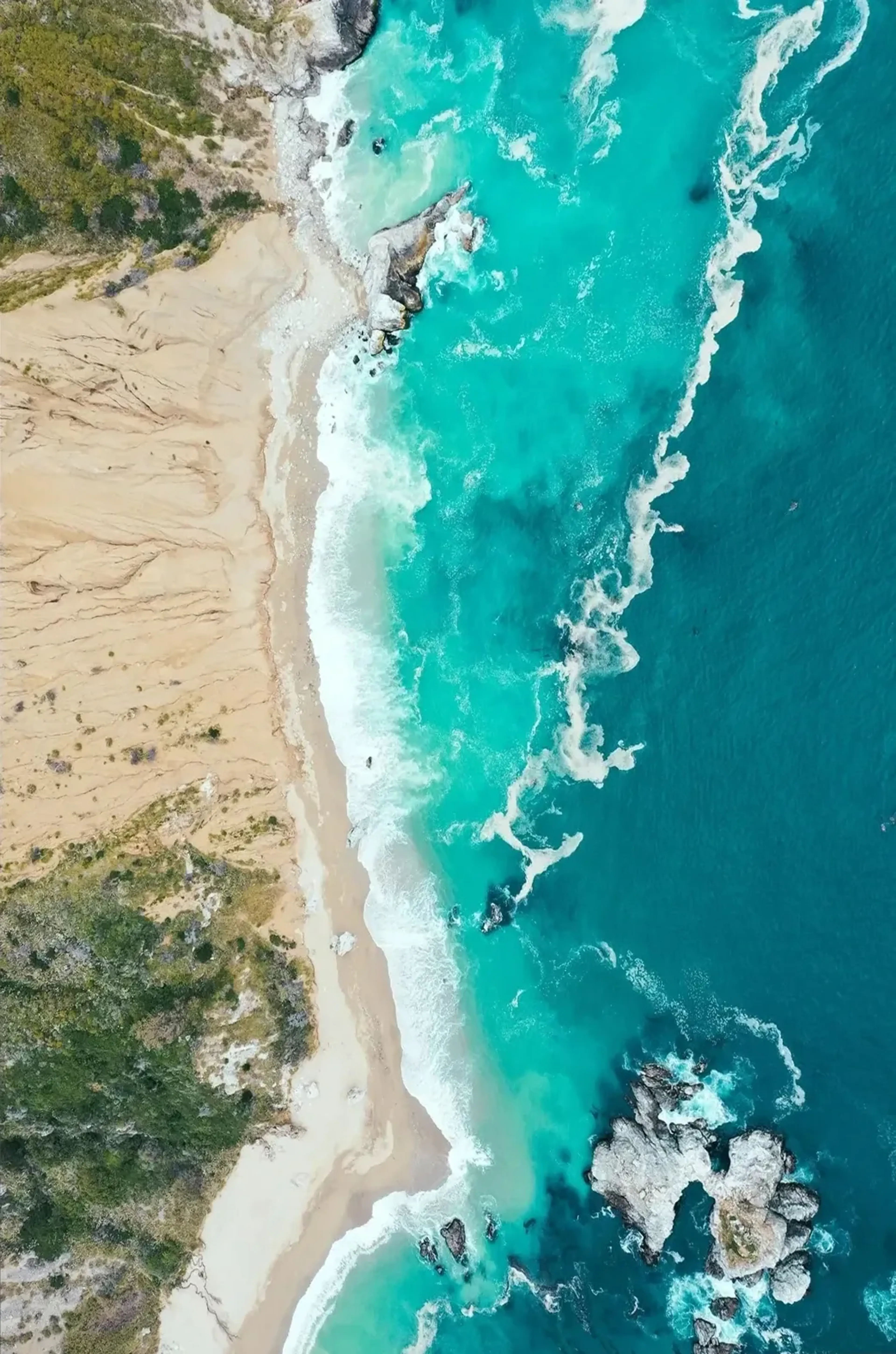 Türkisfarbenes Meer mit weißen Wellen trifft aus der Vogelperspektive auf Sandstrand, Felsen und grüne Vegetation.