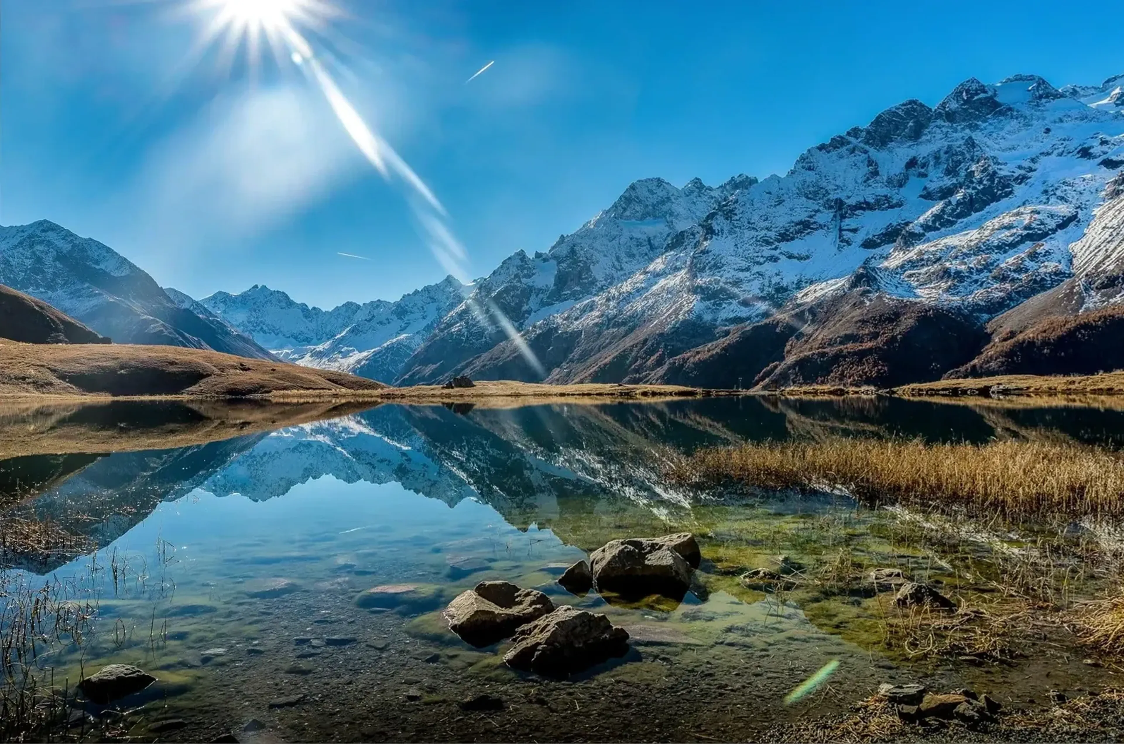 Schneebedeckte Berge spiegeln sich im klaren See, mit Steinen und Gräsern am Ufer unter strahlender Sonne.