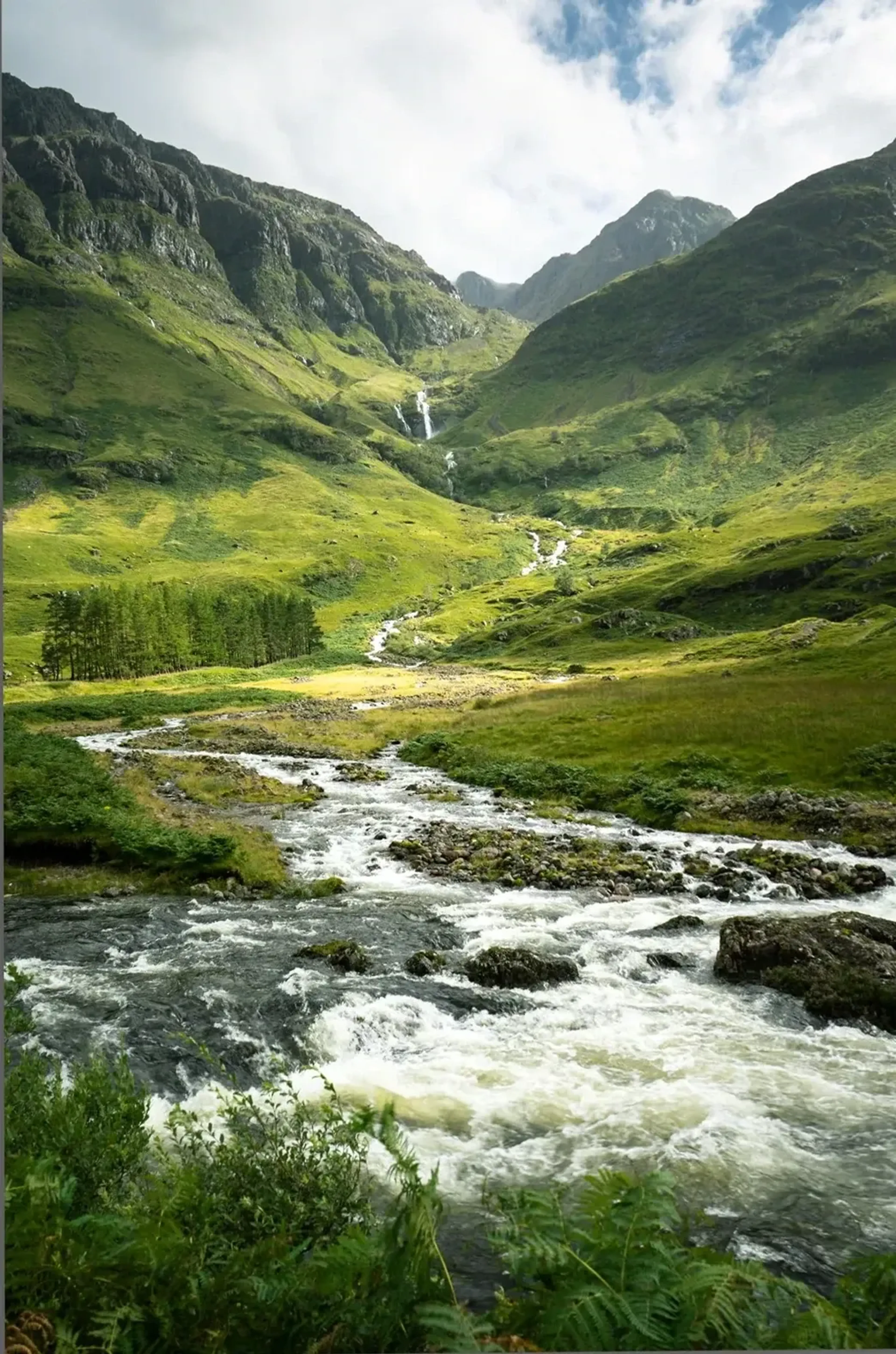 Ein steiniger Bach fließt durch ein grünes Gebirgstal mit steilen Hängen und schmalen Wasserfällen.