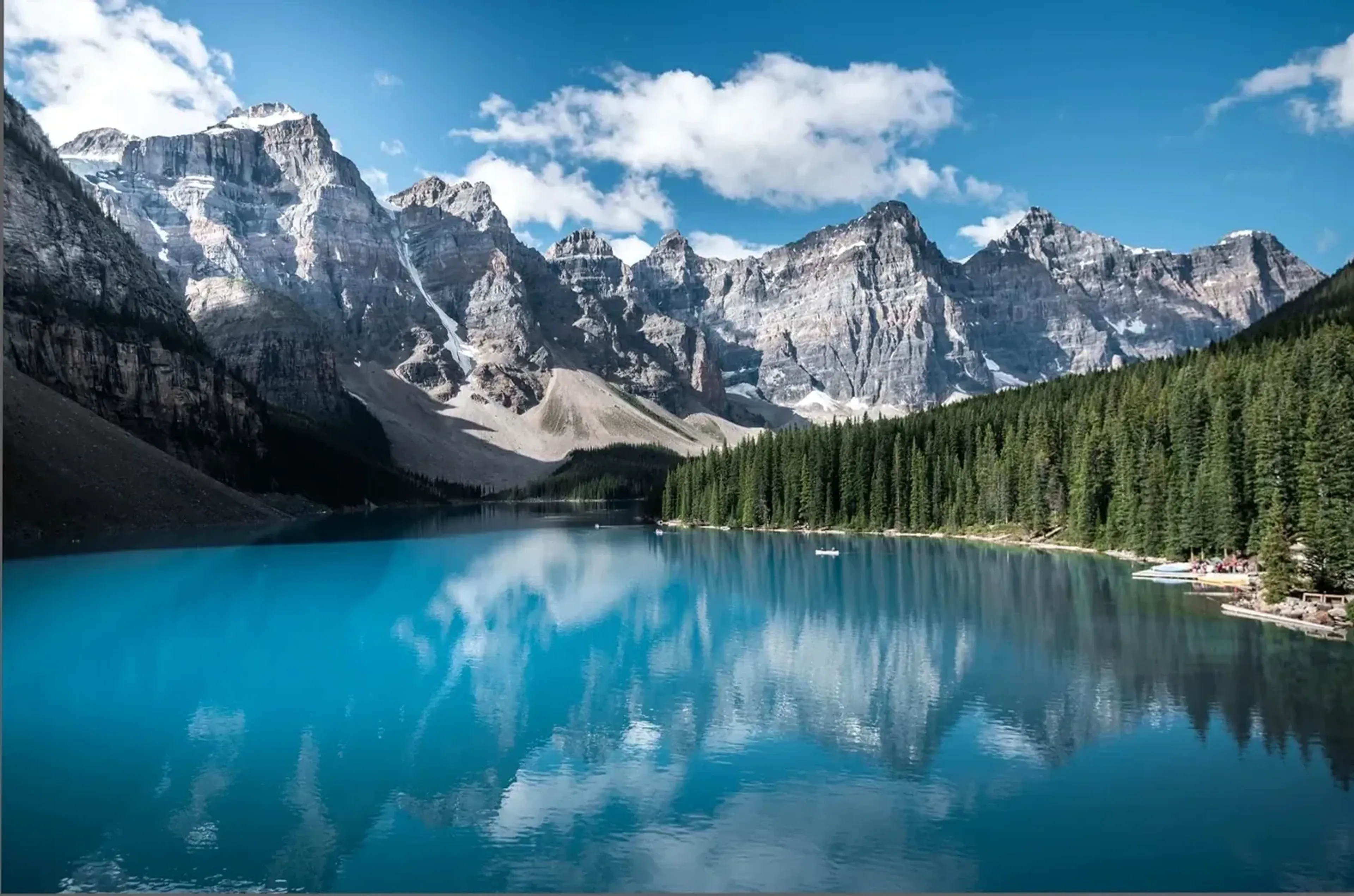 Ein türkisblauer See liegt vor steilen grauen Bergen, mit Nadelwald am Ufer und weißen Wolken am Himmel.