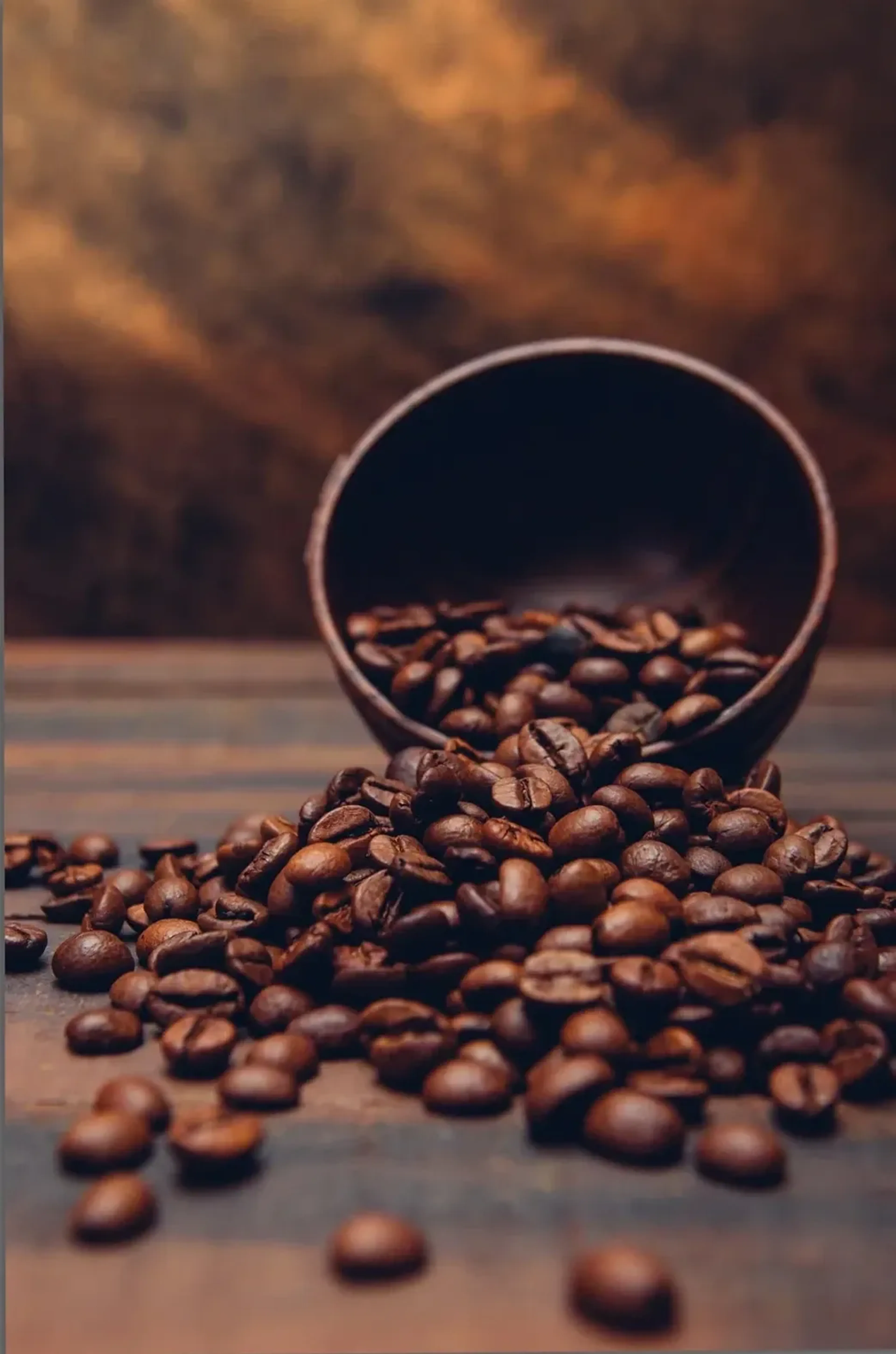 Kaffeebohnen fallen aus einer gekippten dunklen Schale auf eine Tischfläche vor warmbraunem Hintergrund.
