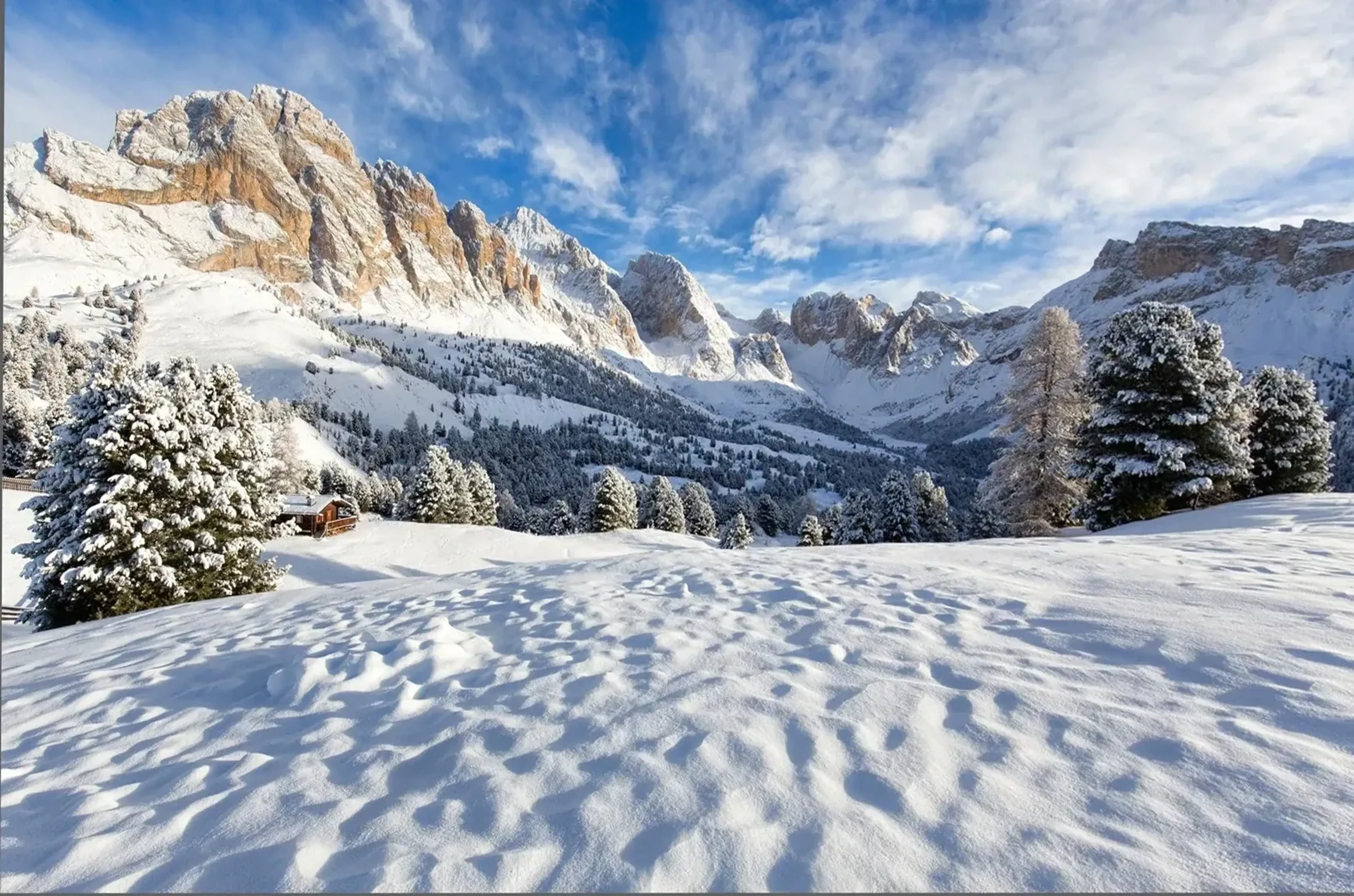Verschneites Bergtal mit Nadelbäumen, kleiner Hütte und hellen Felsgipfeln unter blauem Himmel.