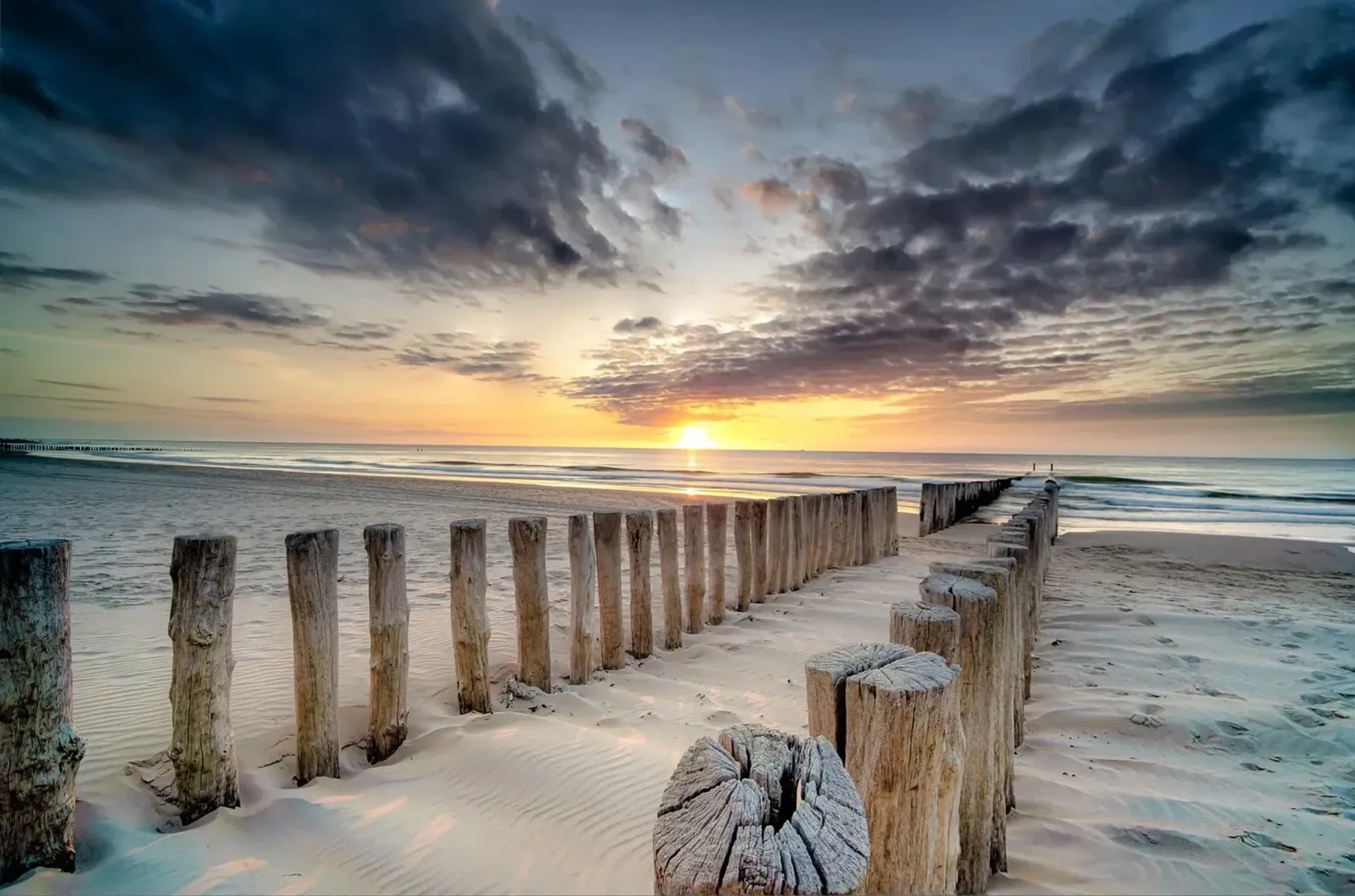 Zwei Reihen verwitterter Holzpfähle führen über einen Sandstrand zum Meer unter einem wolkigen Abendhimmel.