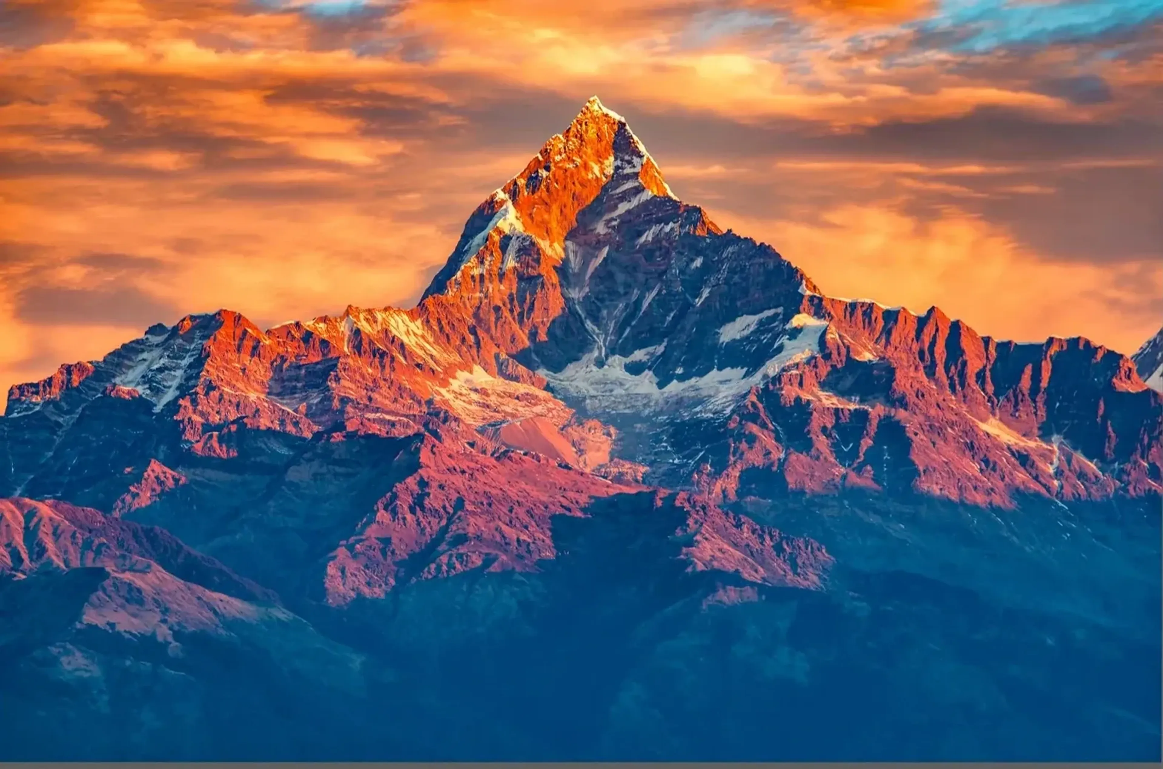 Eine spitze Bergkuppe leuchtet orange über blauvioletten Hängen unter einem rötlich bewölkten Himmel.