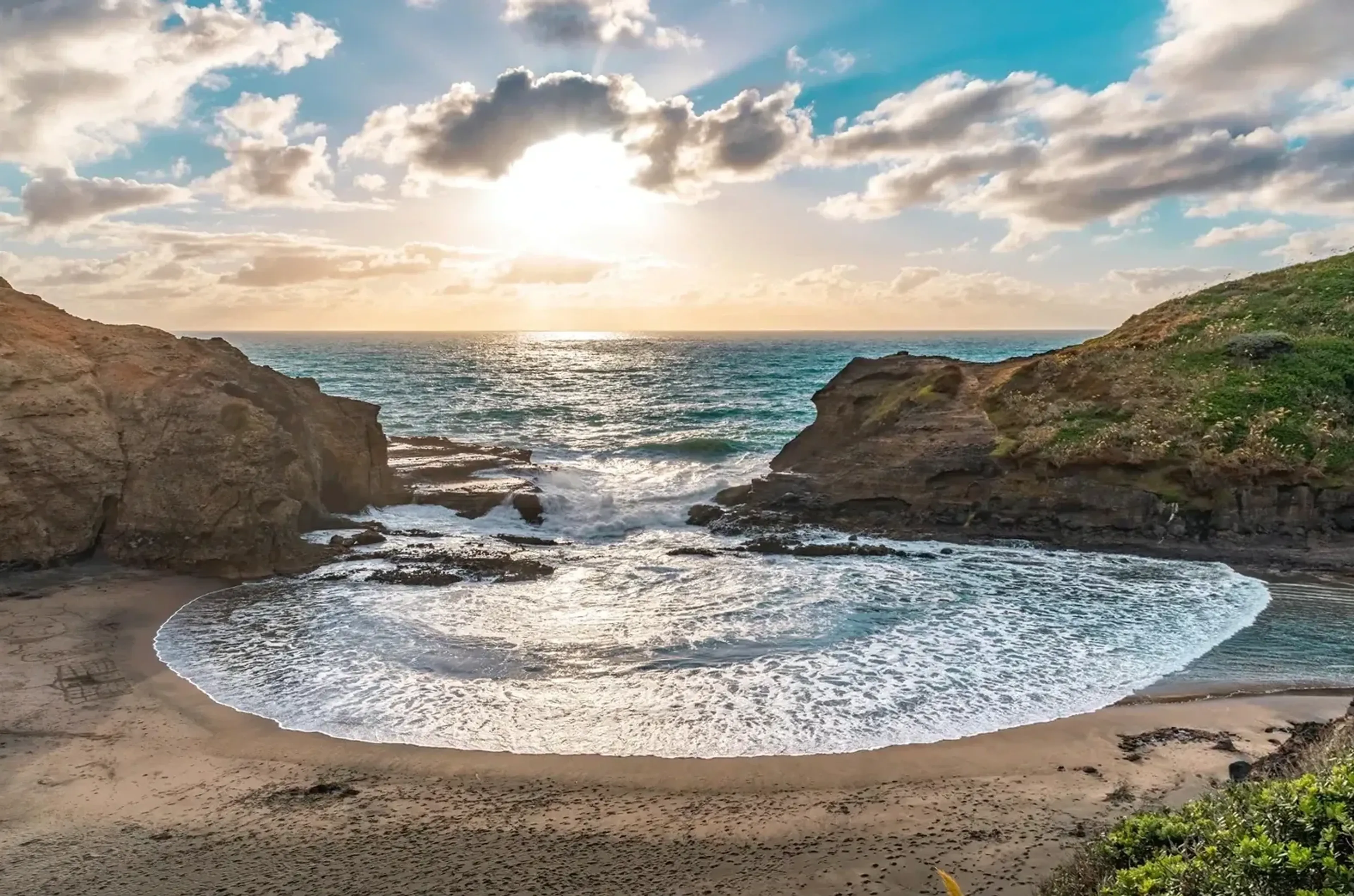 Eine Welle mit weißem Schaum rollt in eine felsige Sandbucht, über der die Sonne durch Wolken scheint.