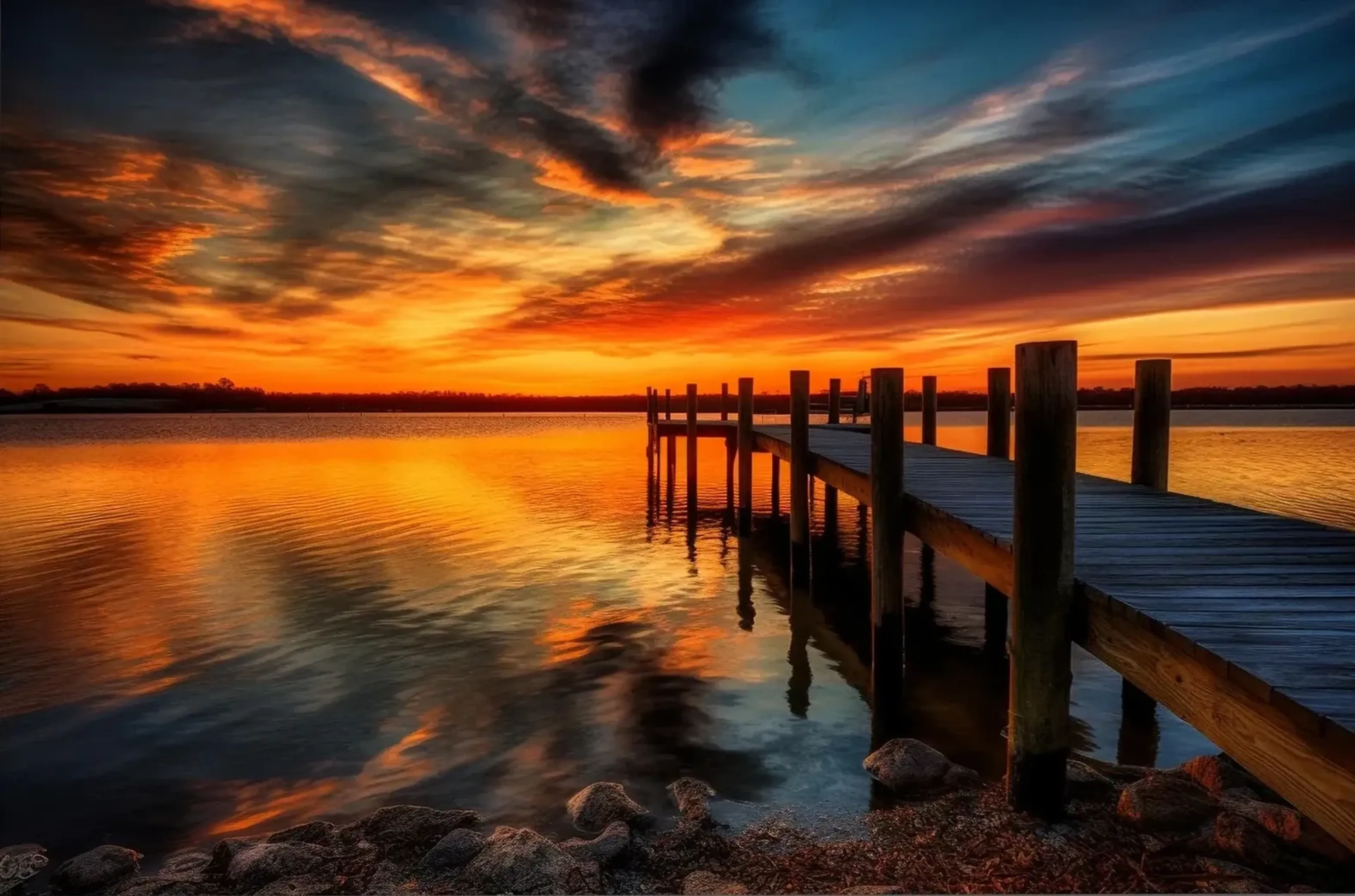 Ein Holzsteg ragt von rechts in einen See, der orangefarbene Abendwolken und dunkelblauen Himmel spiegelt.