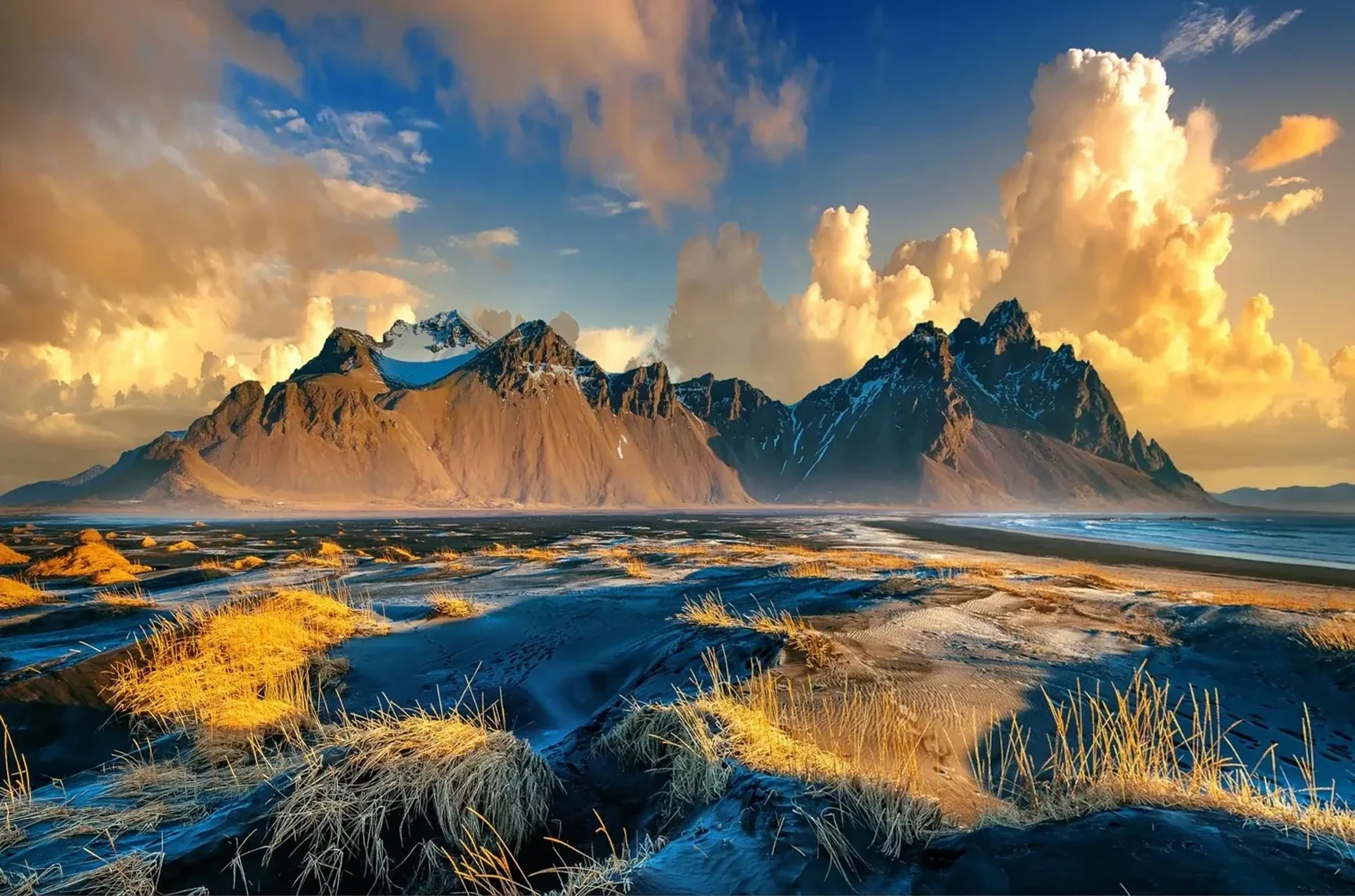 Schroffe Berge hinter dunklen Sanddünen mit goldenen Grasbüscheln, daneben das Meer unter hellen Wolkentürmen.