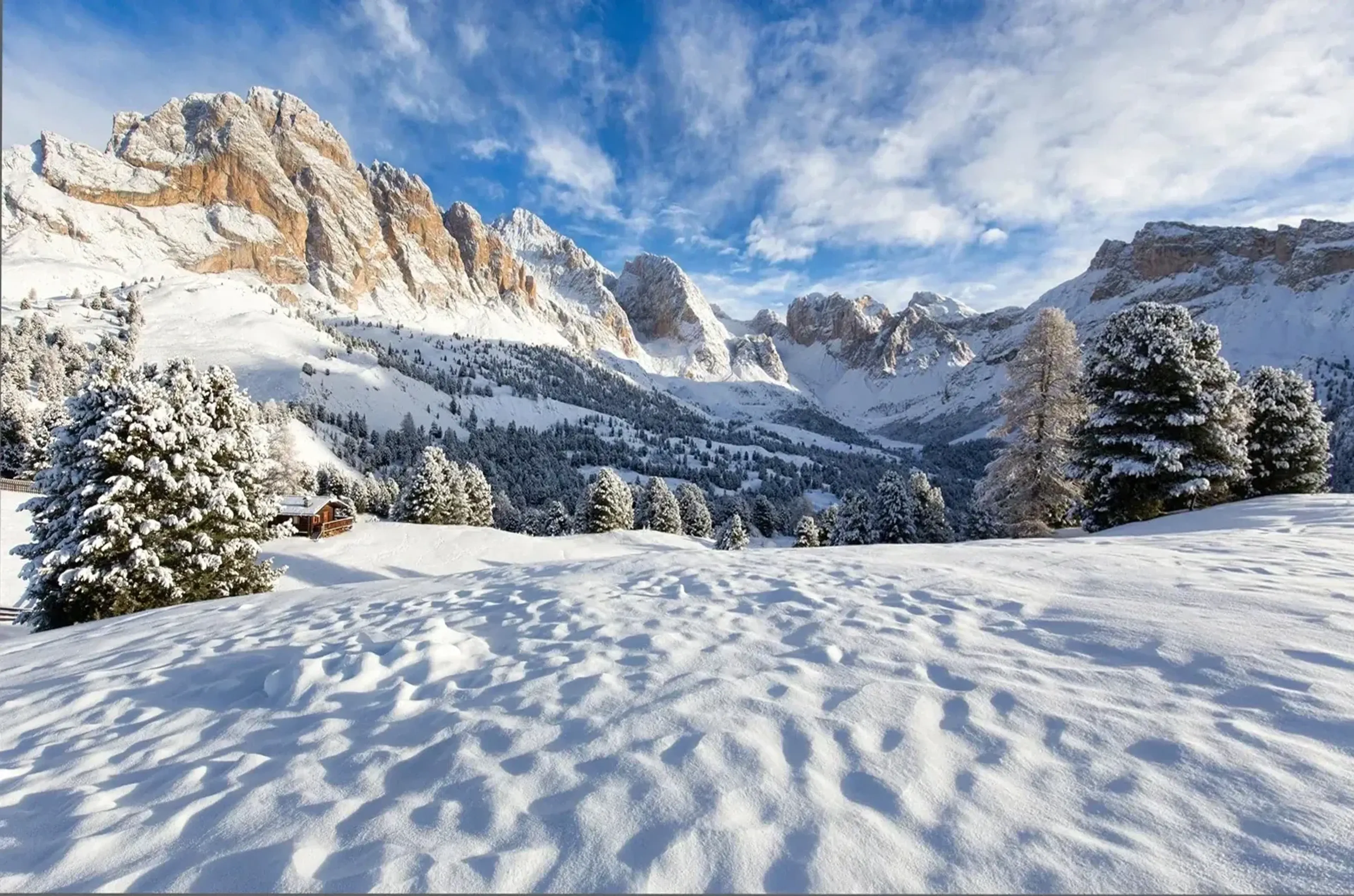 Verschneites Bergtal mit Nadelbäumen, kleiner Hütte und felsigen Gipfeln unter blauem Wolkenhimmel.