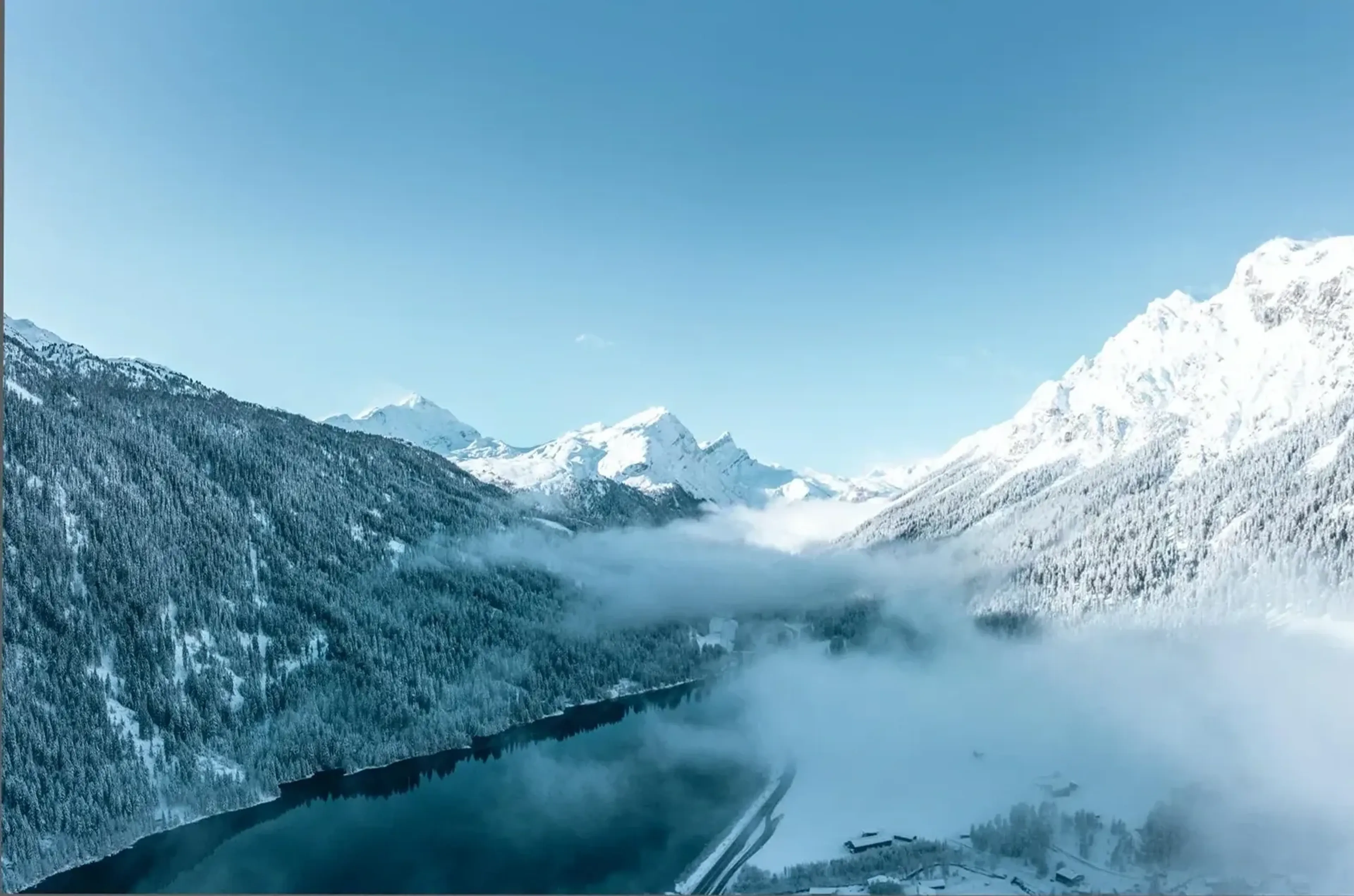 Nebel liegt über einem dunklen See zwischen verschneiten Waldhängen und hellen Berggipfeln.