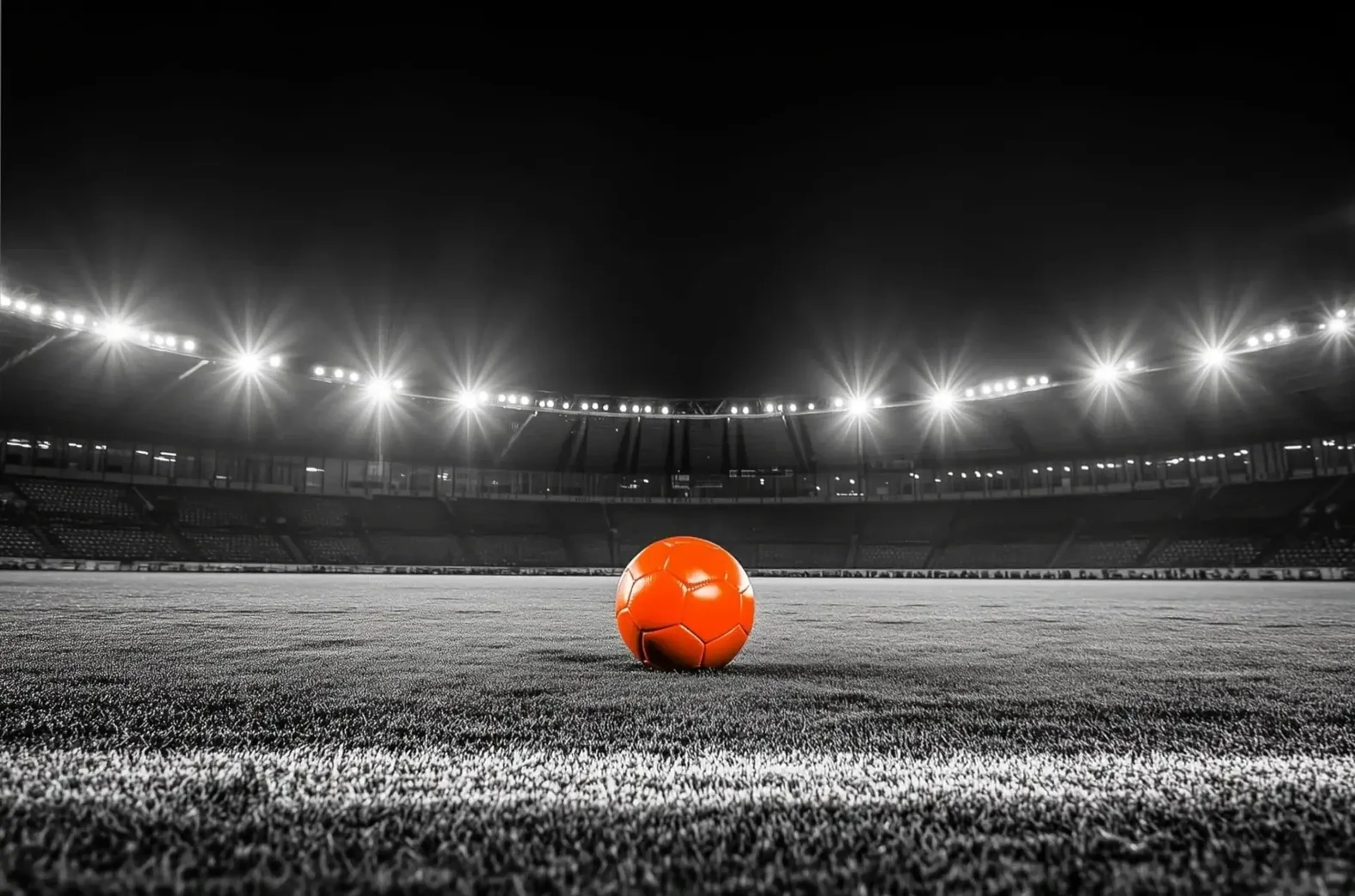 Ein orangefarbener Fußball auf grauem Rasen in einem leeren Stadion unter hellem Flutlicht.