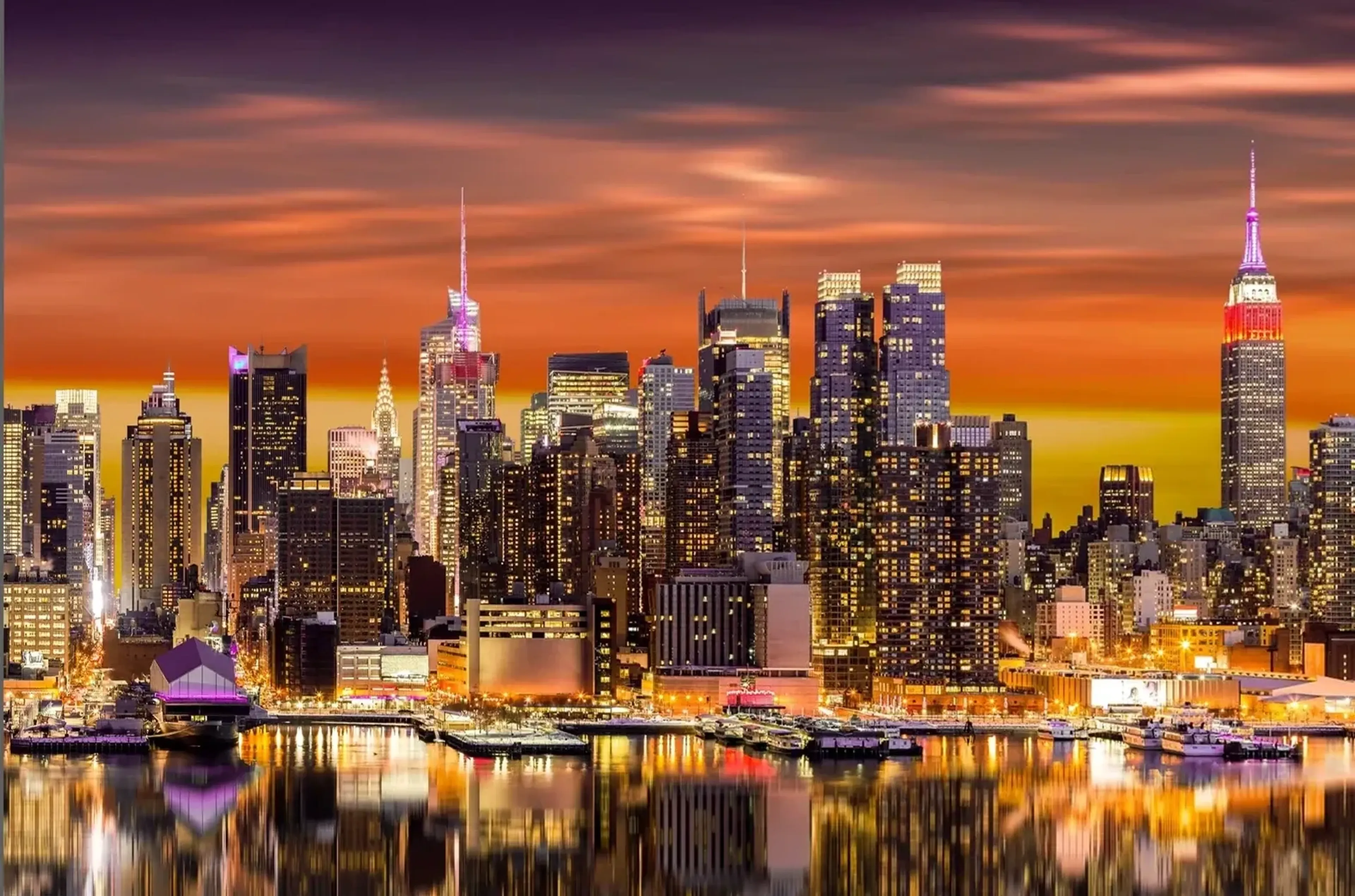 New Yorker Skyline mit beleuchtetem Empire State Building, orangefarbenem Himmel und Lichtspiegelungen im Wasser.