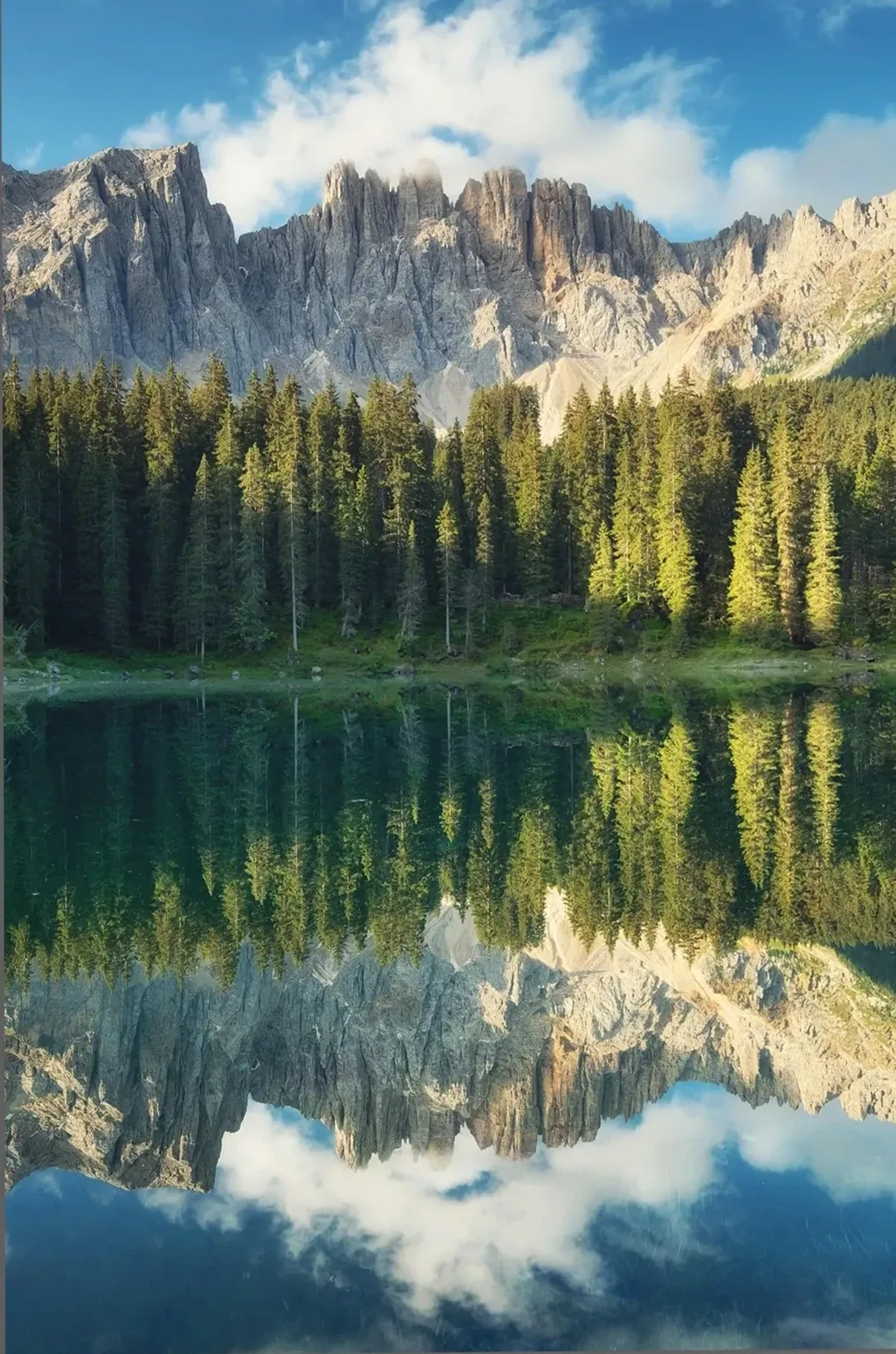 Gezackte Berge, Nadelwald und weiße Wolken spiegeln sich im ruhigen Wasser eines Sees.