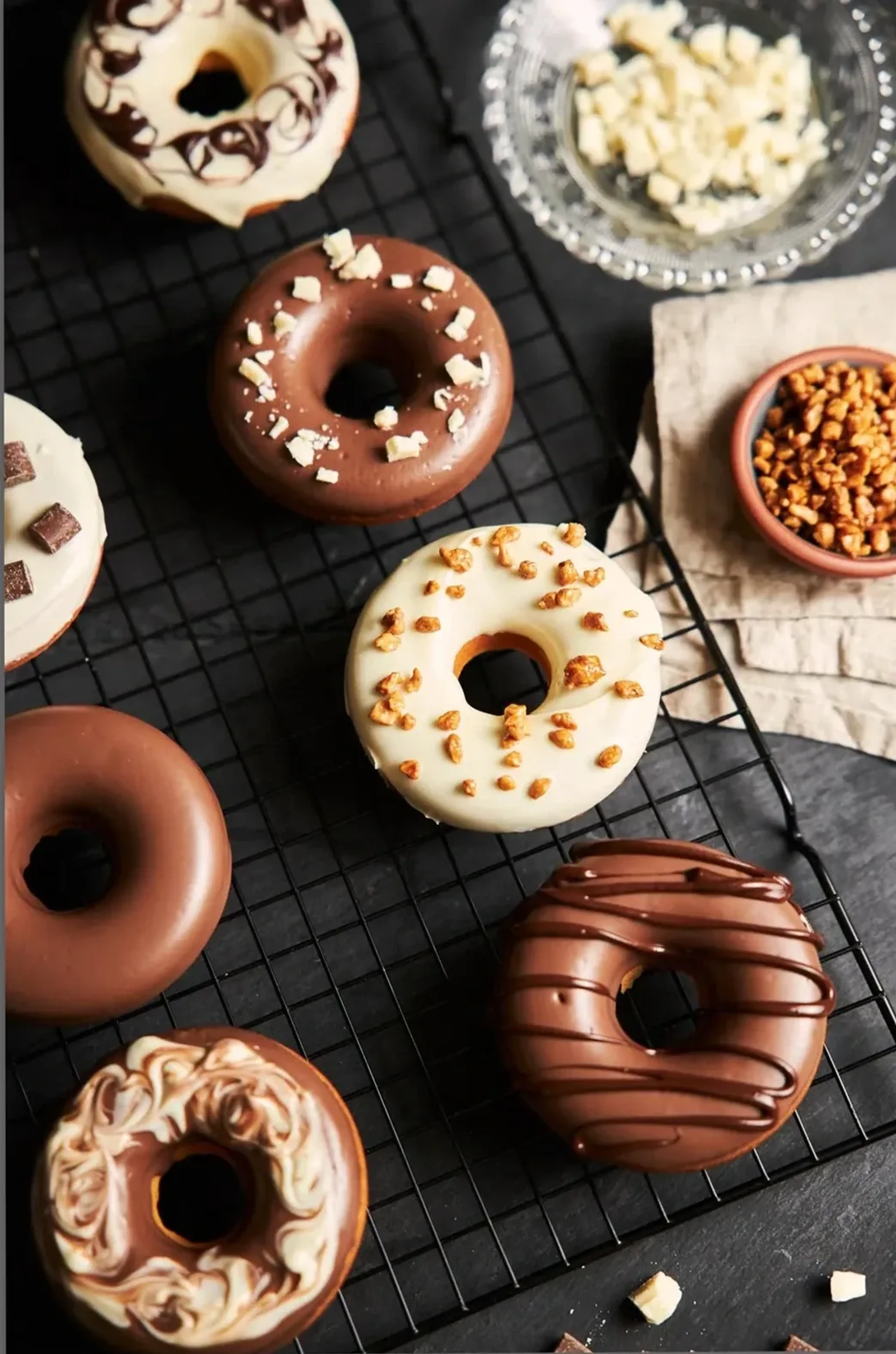 Donuts mit hellen und braunen Glasuren, Streuseln und Schokoladenlinien liegen auf einem schwarzen Gitter.