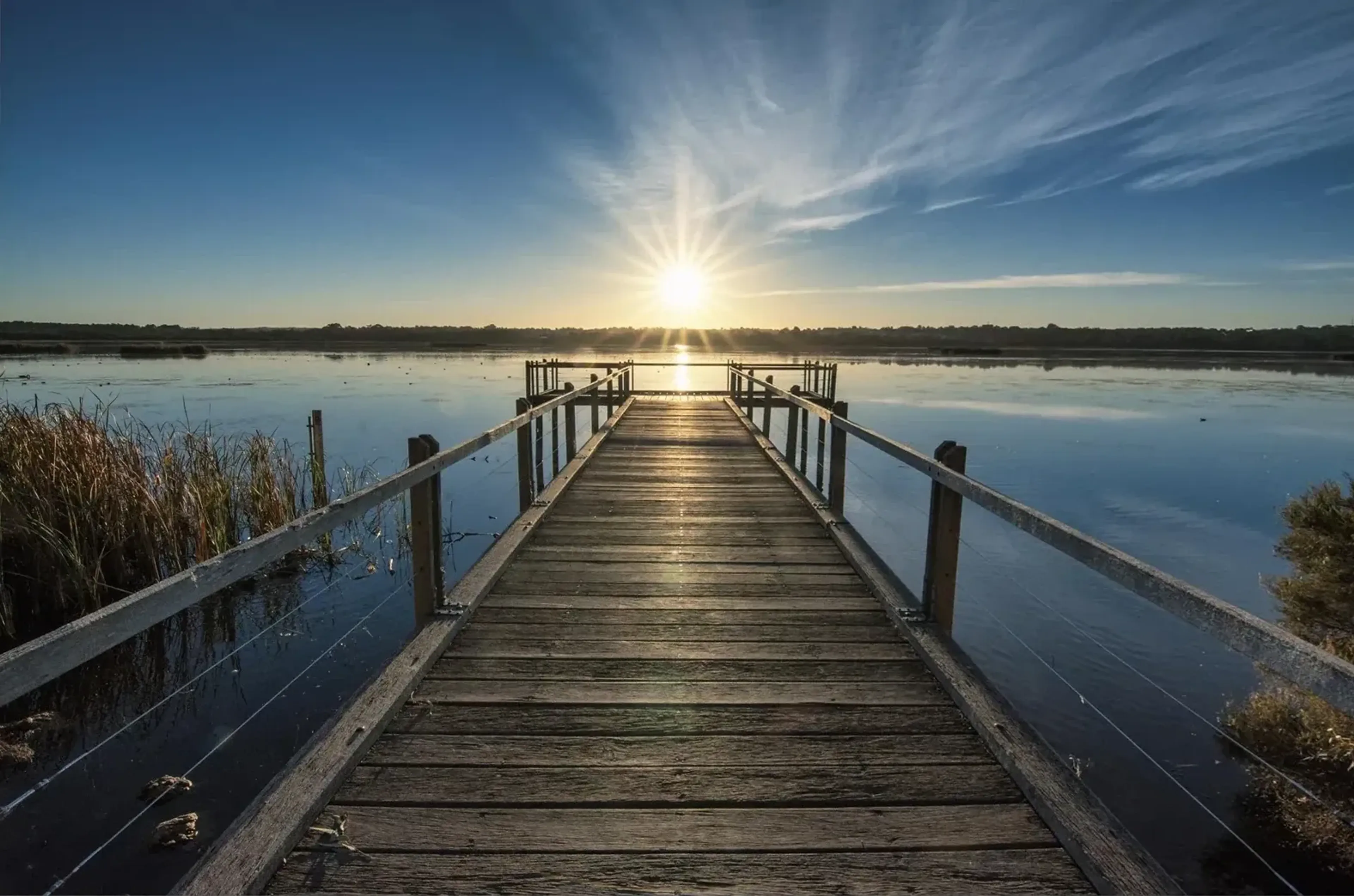 Ein gerader Holzsteg mit Geländern führt über einen stillen See zur tiefstehenden Sonne unter blauem Himmel.