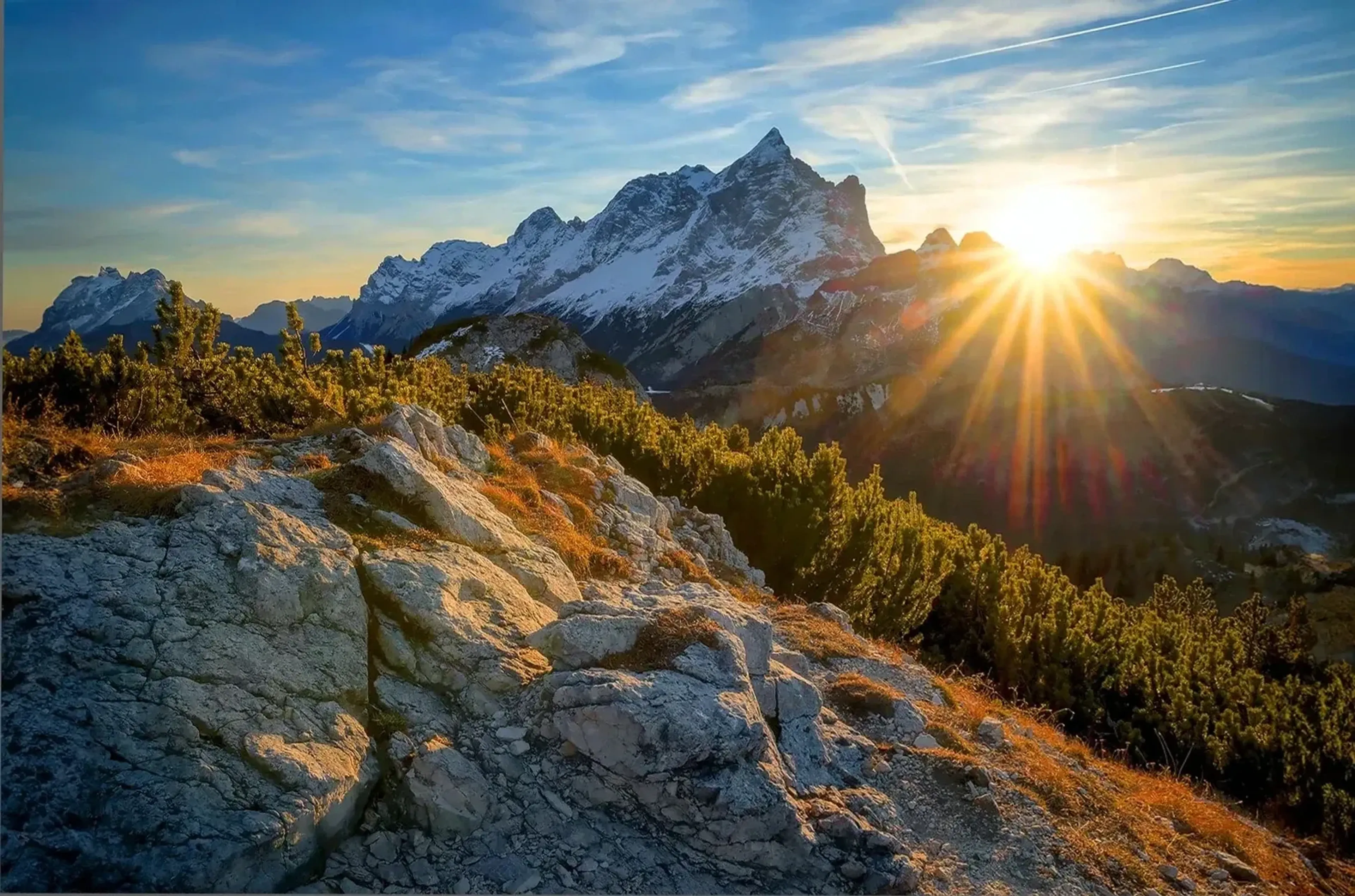 Sonnenstrahlen neben einem schneebedeckten Berggrat, davor helle Felsen und niedrige grüne Sträucher.