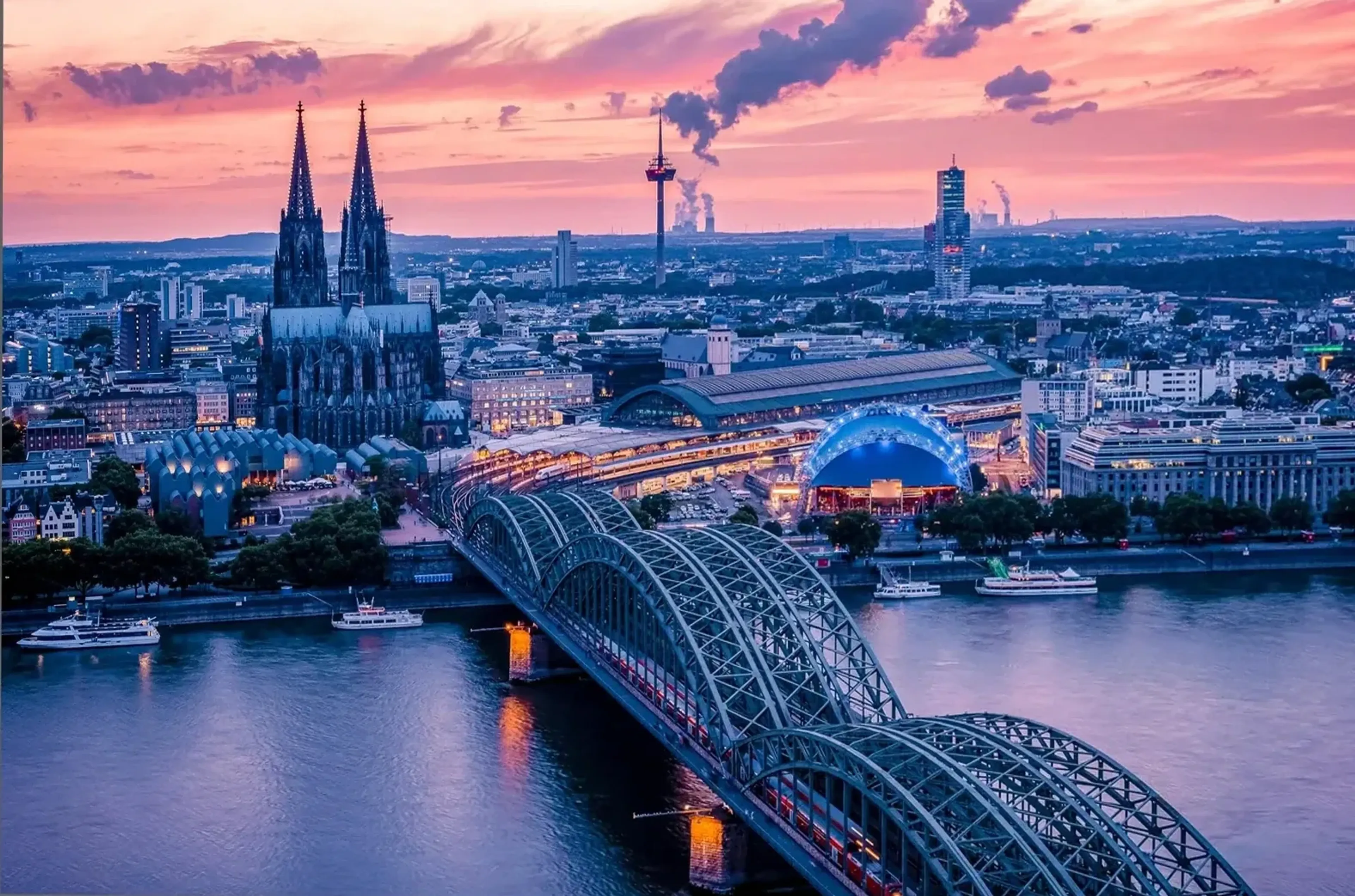 Kölner Dom, Hauptbahnhof und Hohenzollernbrücke am Rhein unter einem rosa-violetten Abendhimmel.