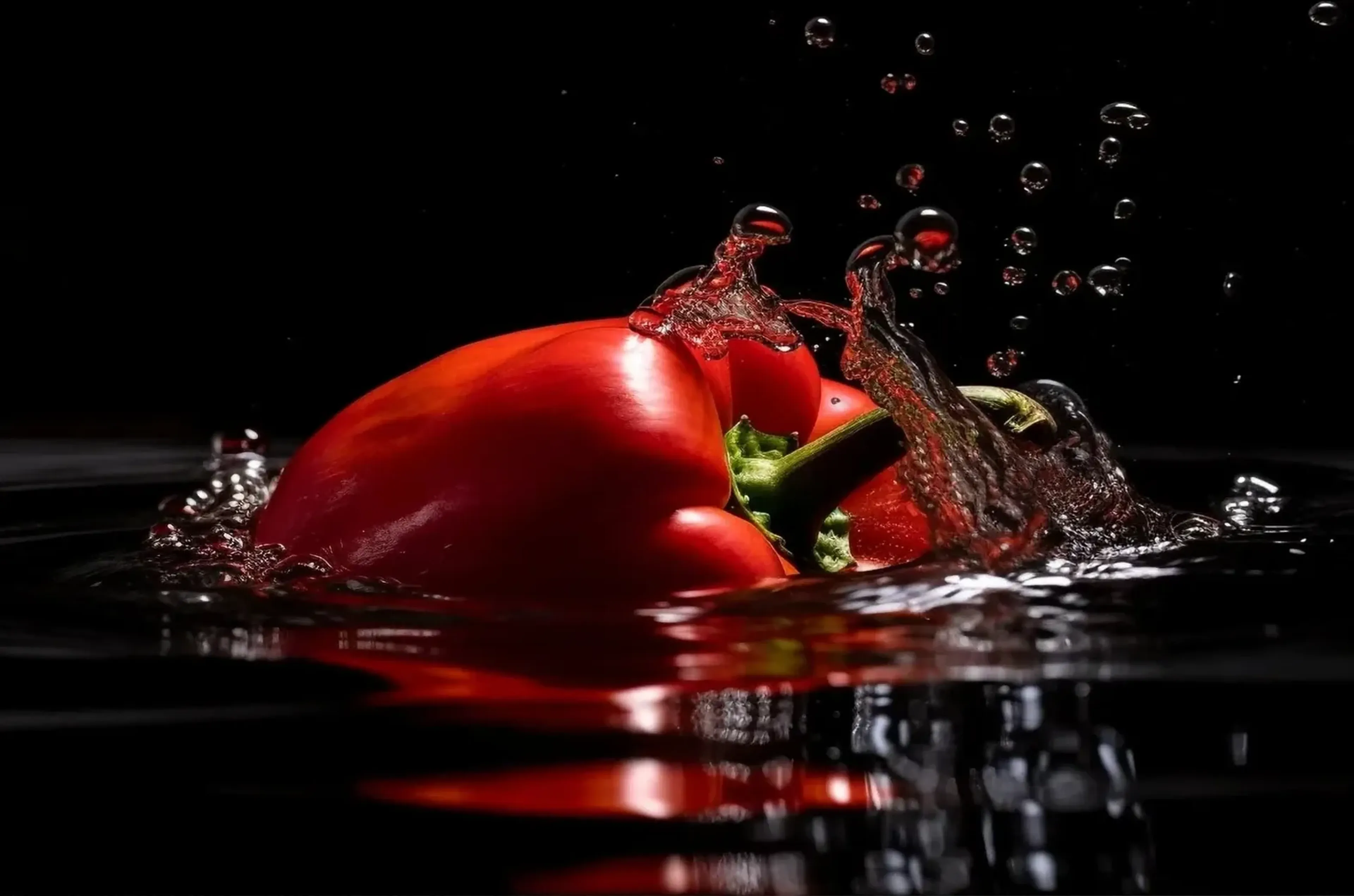 Eine rote Paprika mit grünem Stiel liegt zwischen Wasserspritzern und Tropfen auf einer dunklen Wasserfläche.
