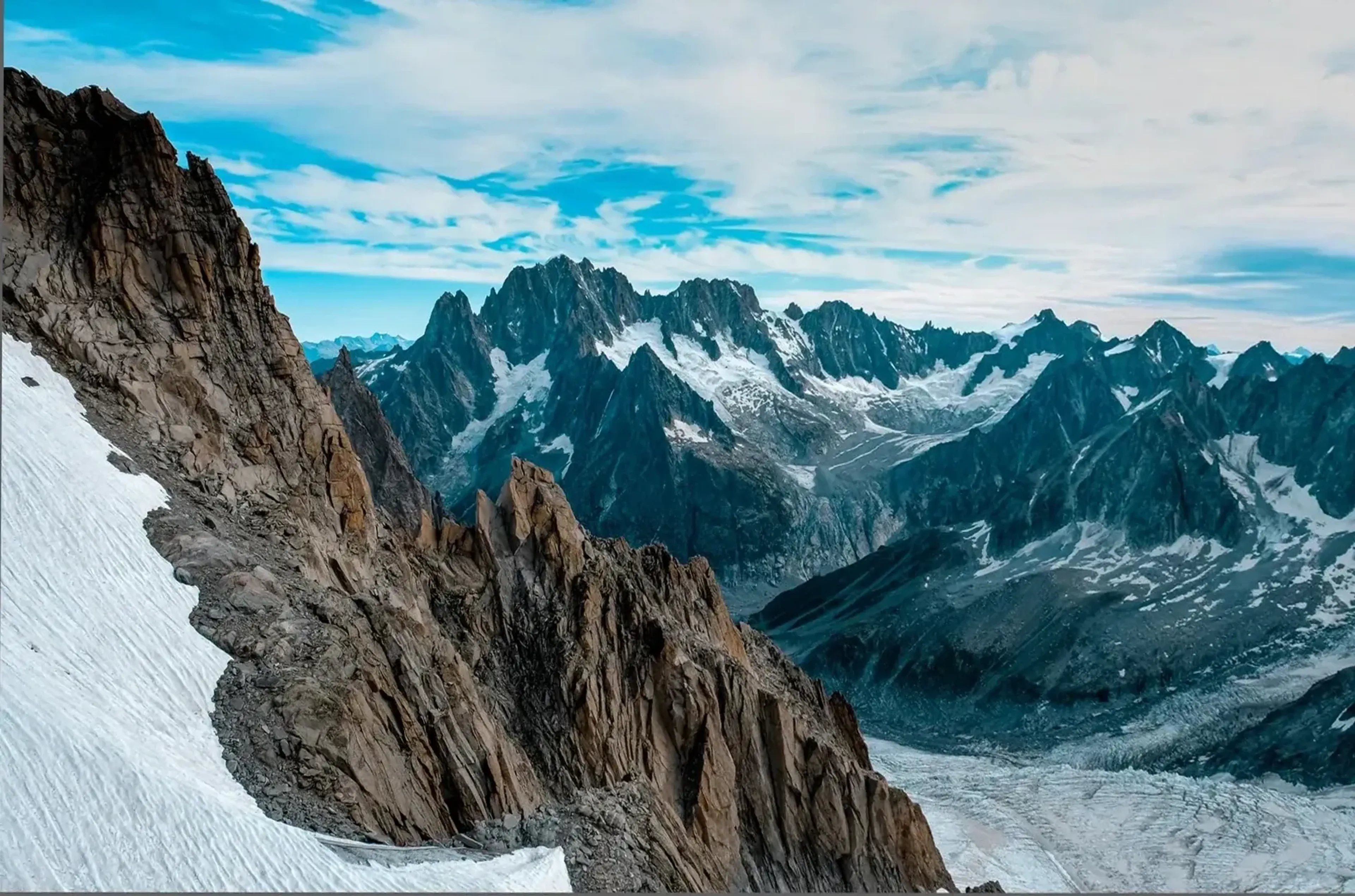 Eine schroffe Felswand und ein Schneefeld liegen vor einem breiten Gletscher und gezackten Bergen unter blauem Himmel.