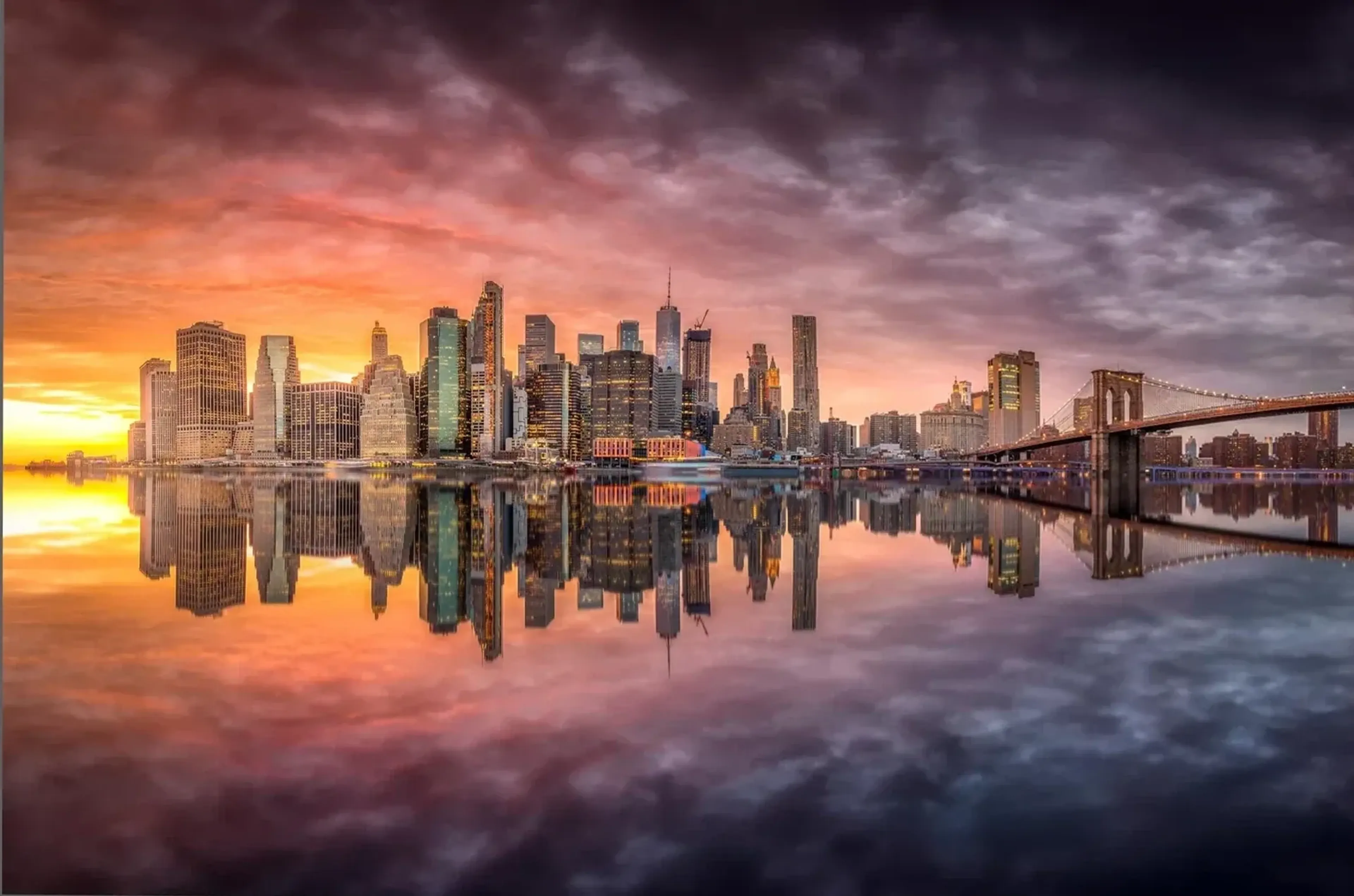 New Yorker Skyline und Brücke spiegeln sich unter orangefarbenen und violetten Wolken im Wasser.